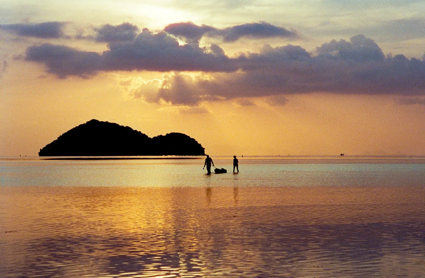 Two people wading in shallow ocean water during a sunset, holding a bag or object between them with an island in the background and clouds in the sky.