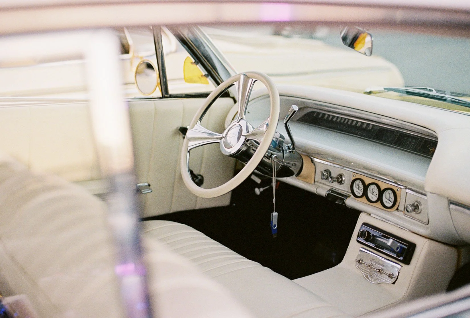 Interior view of a vintage car showing a white steering wheel, dashboard with gauges, and cream-colored upholstery.