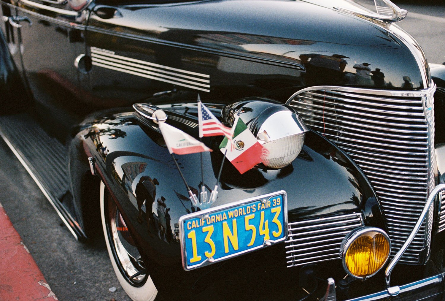 Close-up of the front of a vintage black car with a California license plate that reads "13N54X3". The car has small American, Mexican, and Italian flags mounted on the hood, along with a chrome headlight, yellow fog light, and chrome grille. Reflect