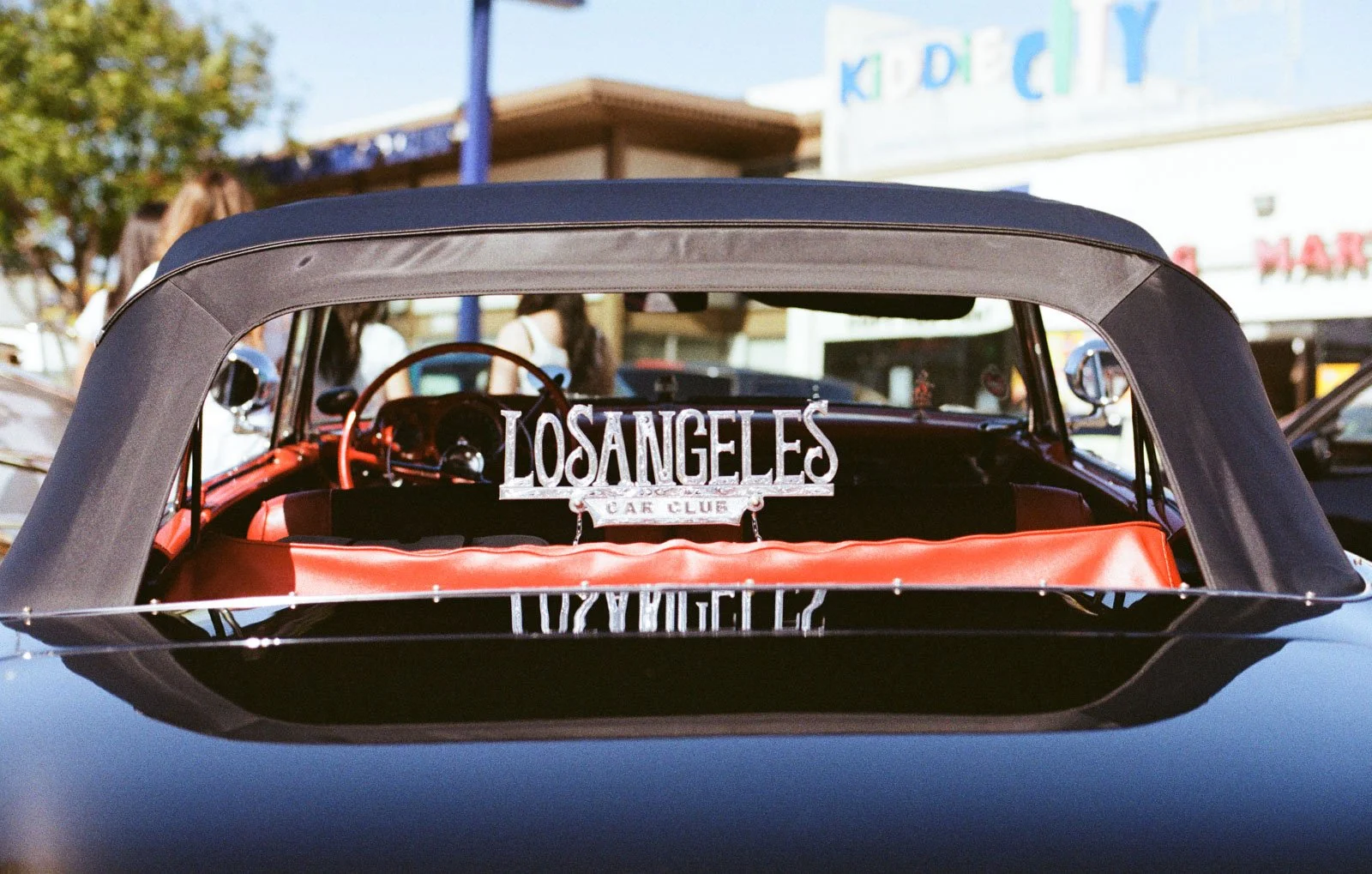 The interior of a classic black convertible car with a Los Angeles Car Club sign visible through the rear window, parked outside in front of a building with a sign that reads Kid City.