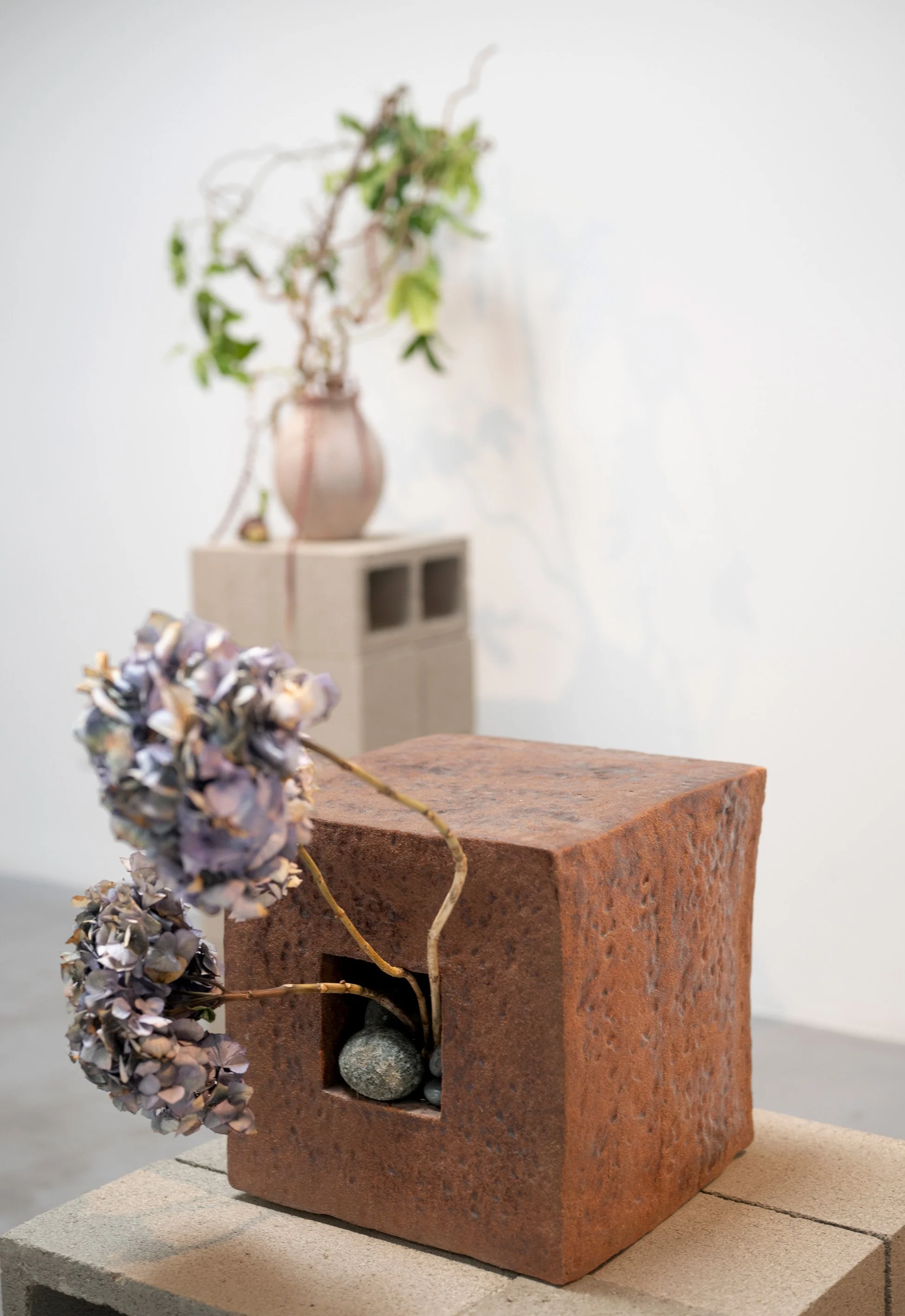 A decorative arrangement featuring a pinkish ceramic vase with greenery, dried flowers, and a small black stone, placed on a brick block against a neutral background.