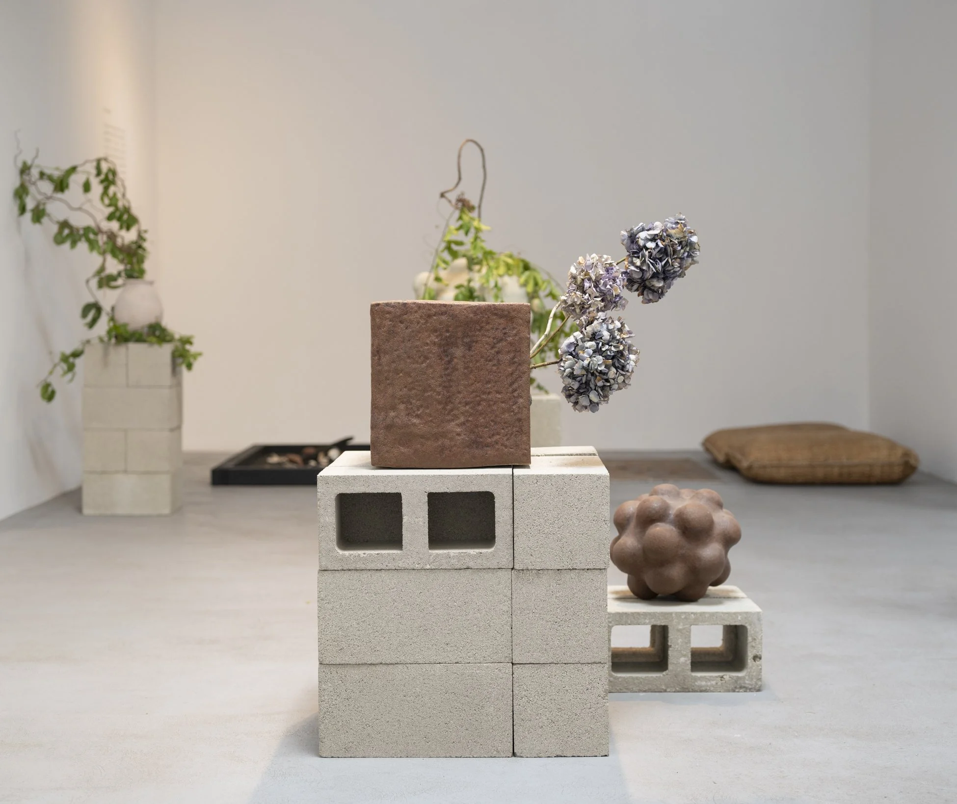 Display of various items including a cinder block plinth with a vine plant, a botanical sculpture made of hydrangeas, and a brown cushion on the floor next to a grey rug