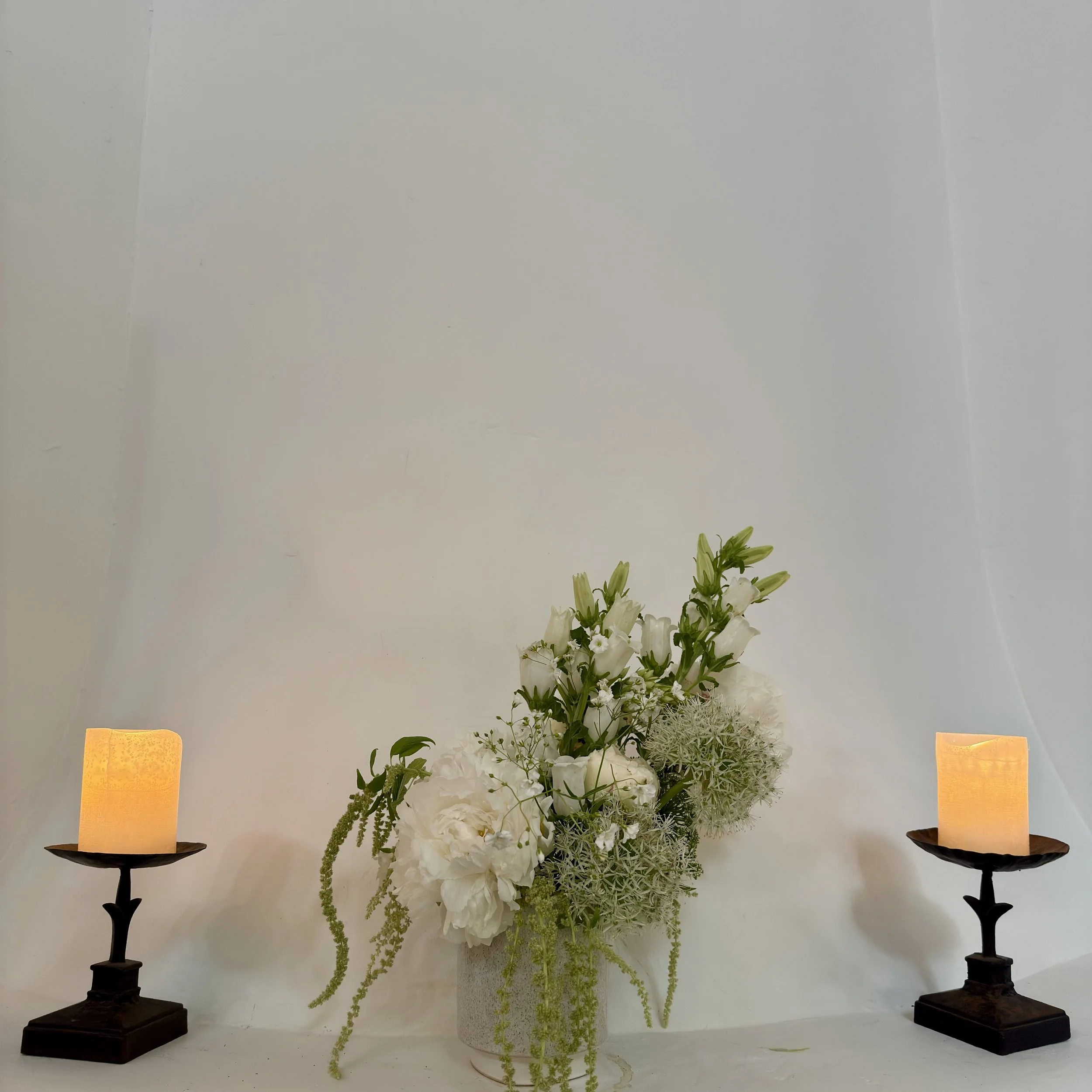 A floral arrangement in a gray pot with white flowers and greenery, flanked by two lit candles on dark candleholders, against a plain white background.