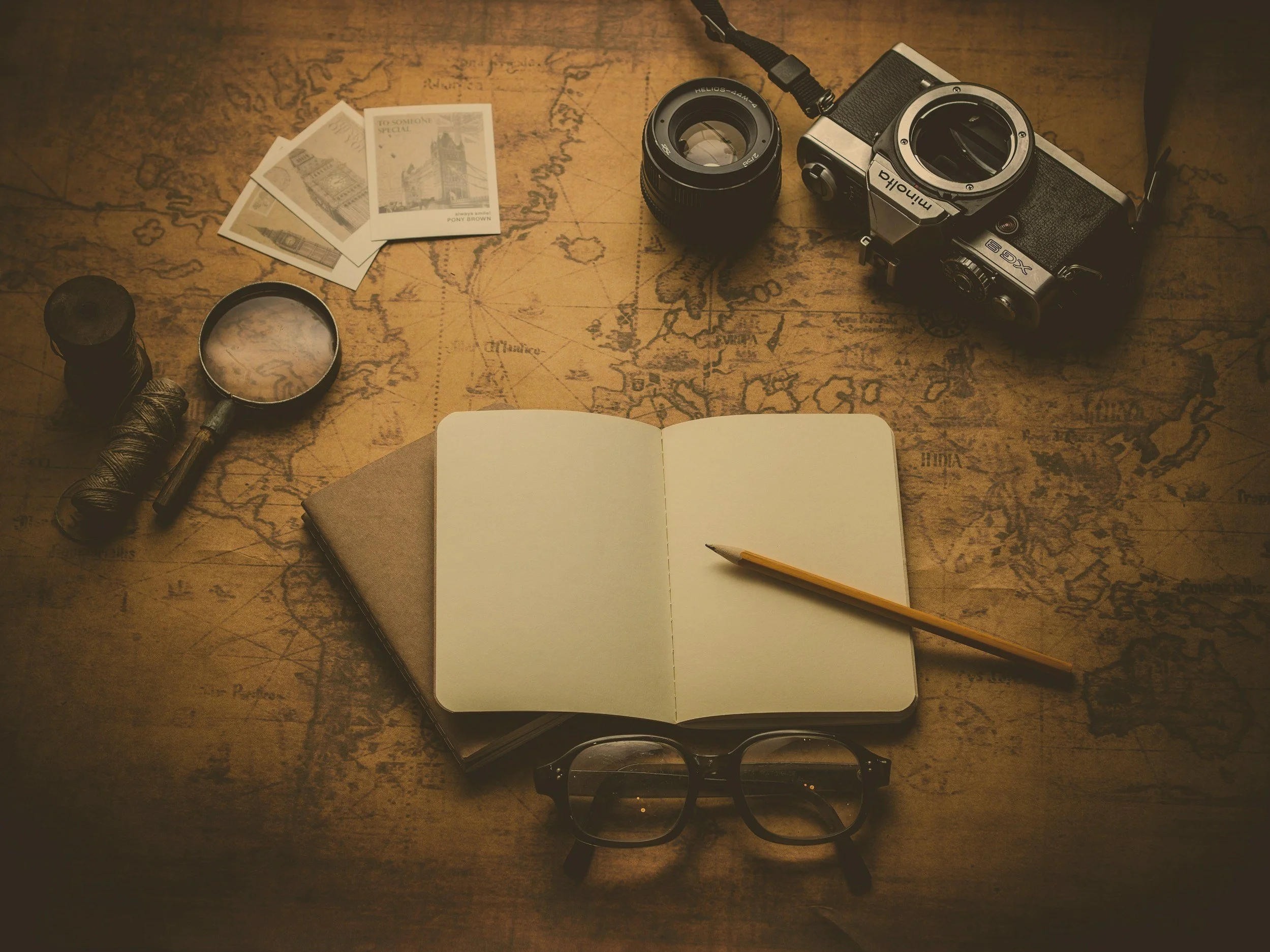 Vintage map with a camera, lens, photographs, magnifying glass, notebook, pencil, glasses, and a spool of thread arranged on it.