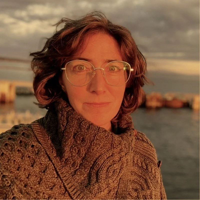A woman with curly short hair and glasses near a body of water during sunset.