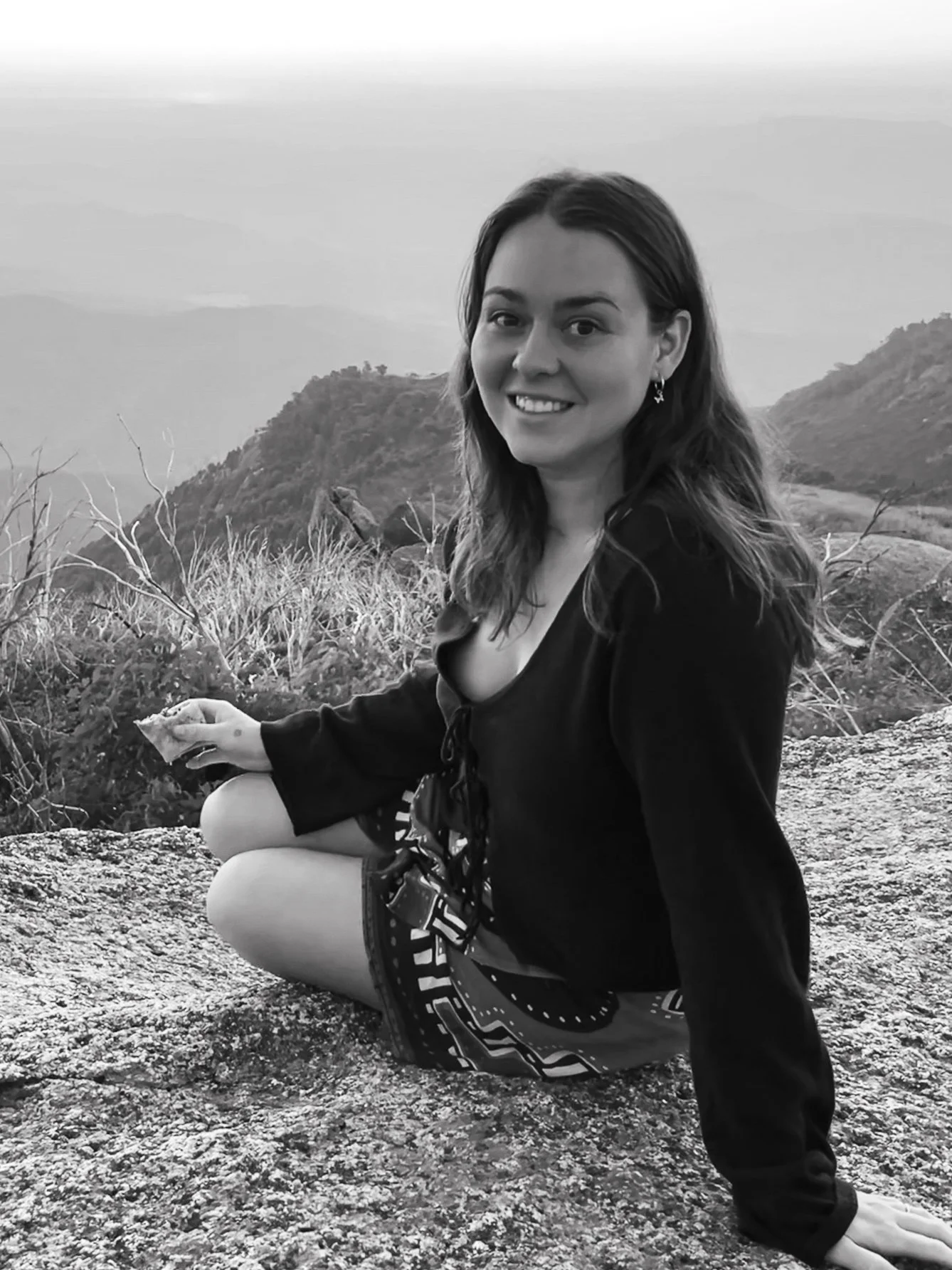 Smiling woman sitting on a rock with mountain view in the background, black and white photo.