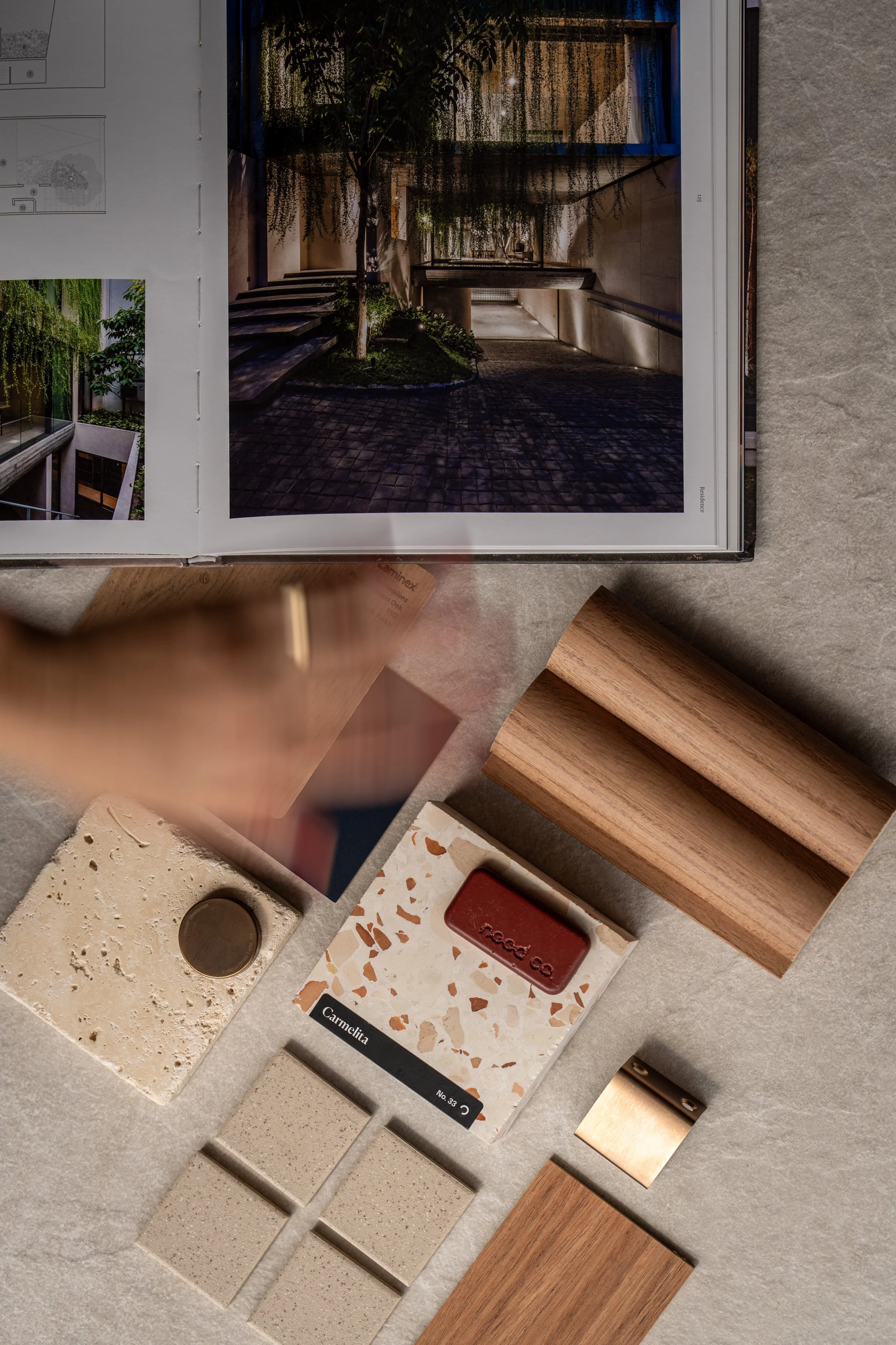 Top view of various architectural and interior design materials, including wood, stone, and metal samples, placed on a textured surface next to an open architecture book displaying outdoor spaces.