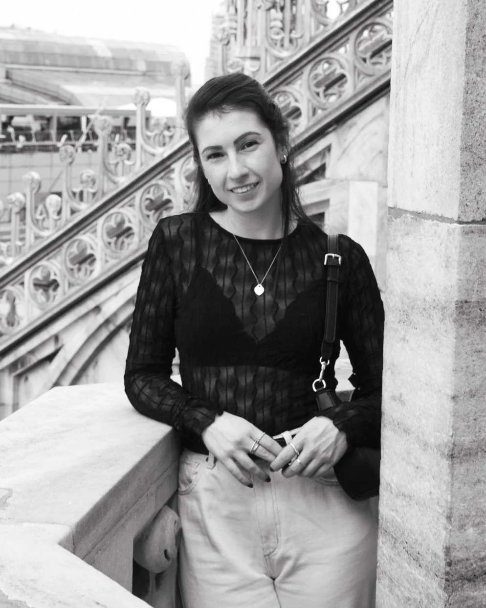 Woman in black lace top and light-colored pants leaning on a stone railing, with ornate architectural details in the background.
