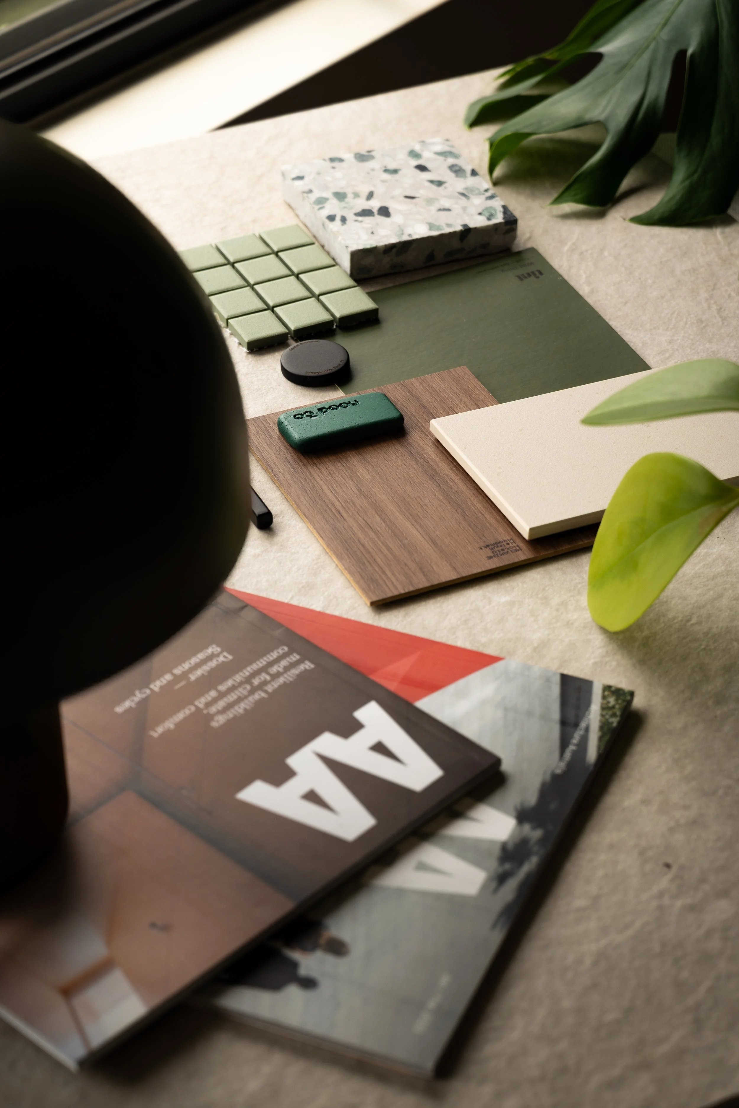 Interior design mood board with green and brown color swatches, magazines, and plant leaves on a table.