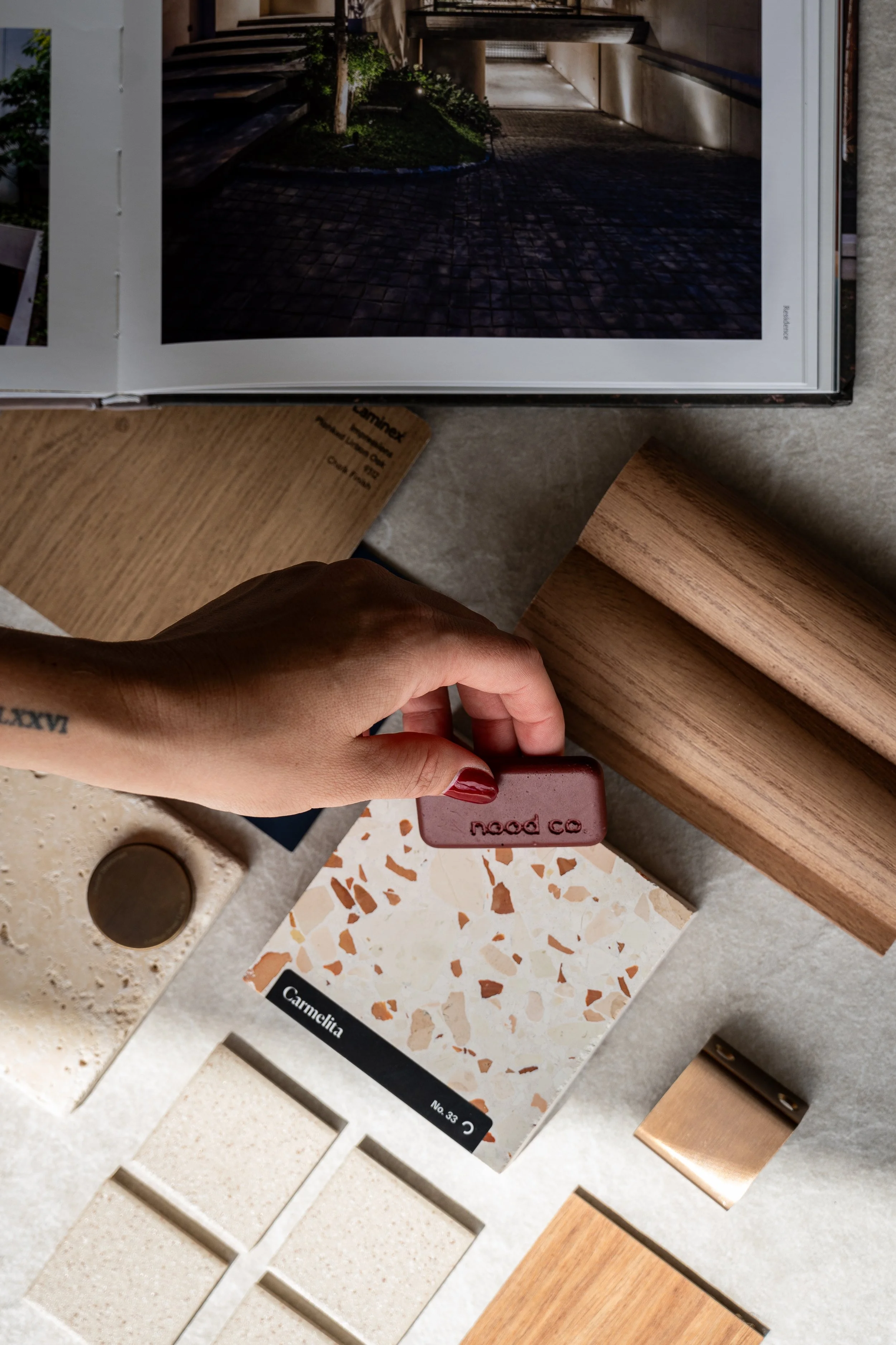 A person holding a small terracotta-colored sample with the word 'nod co.' embossed on it, surrounded by various terrazzo, wood, and stone samples on a table.