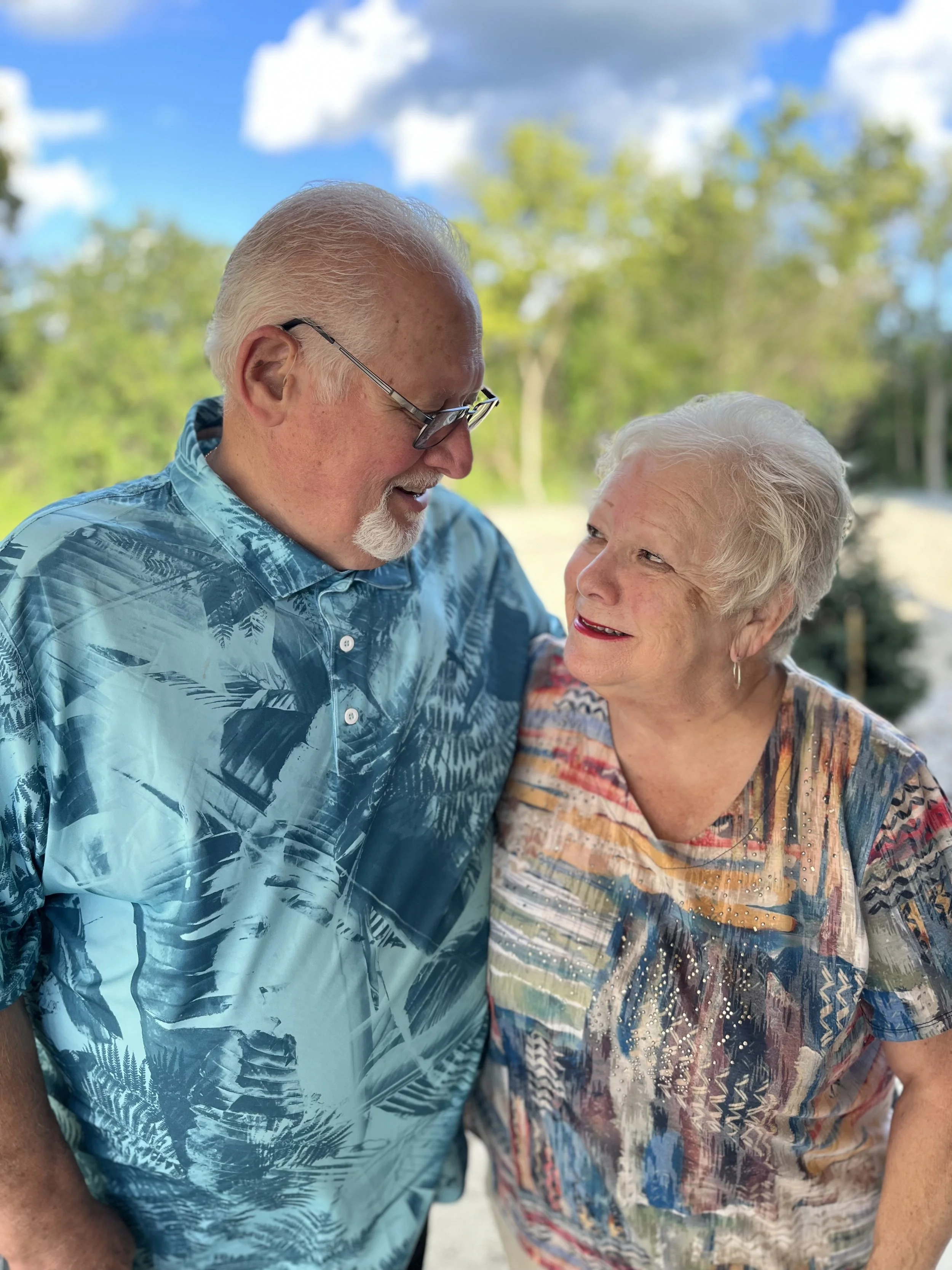 A close-up photo of an elderly man with gray hair and glasses smiling and looking at an elderly woman with gray hair and red lipstick as they stand outdoors on a sunny day with trees and blue sky in the background.