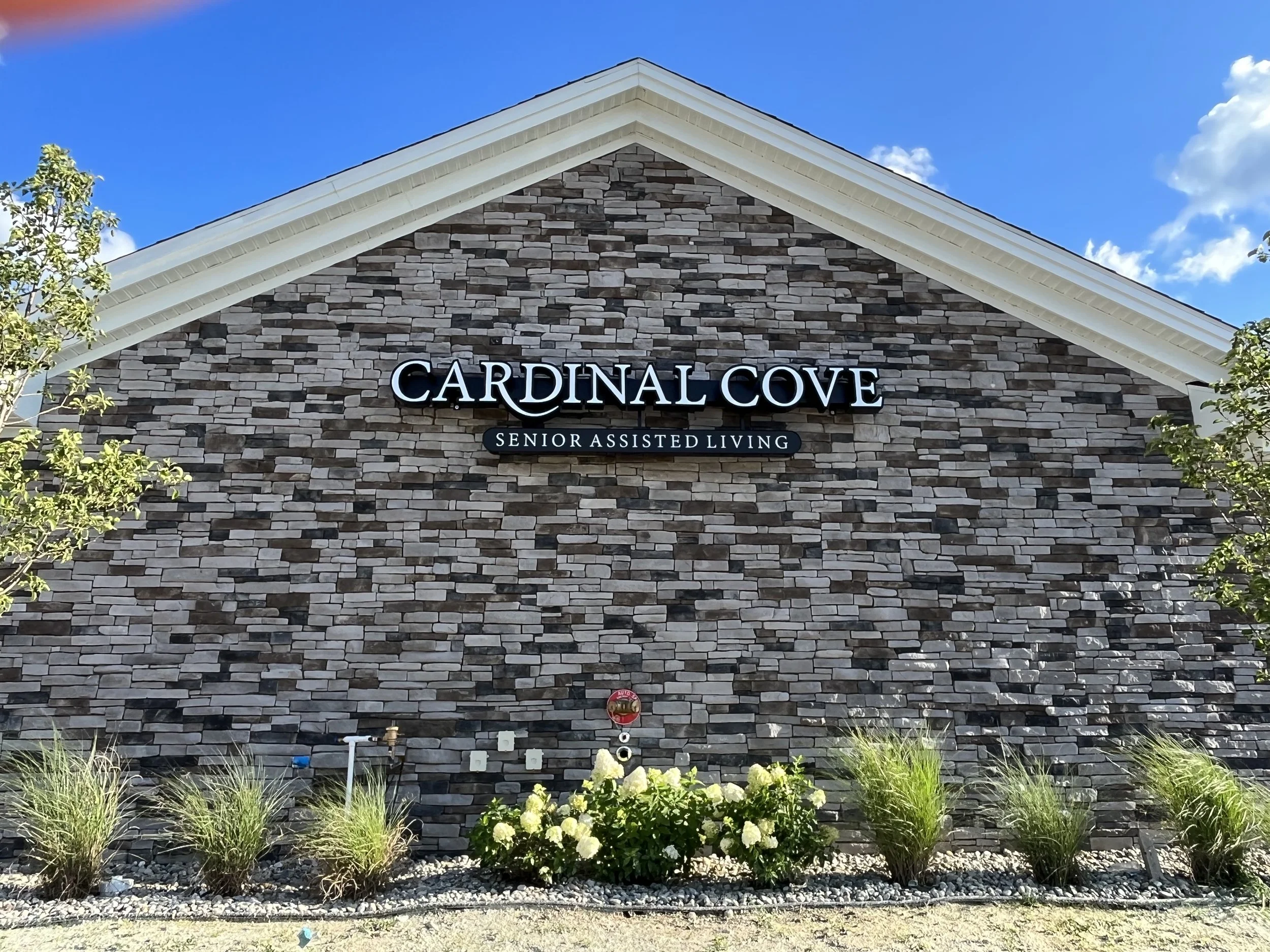 Front view of a building with a stone facade and a sloped roof, signage reads 'Cardinal Cove Senior Assisted Living', with landscaping in the foreground and cloudy blue sky above.