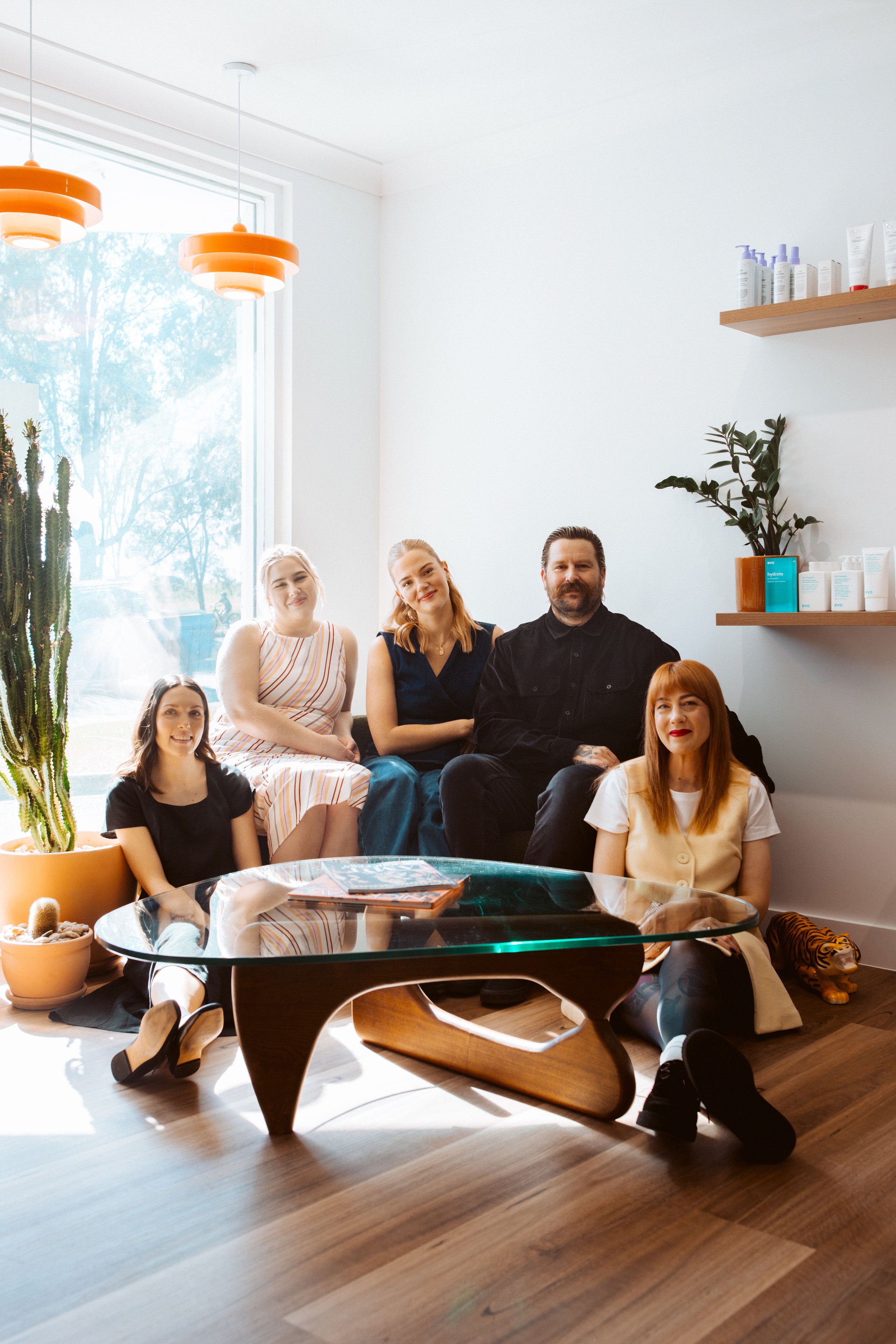 Group of five people sitting in a modern, well-lit room with a large window, wooden floors, a cactus plant, and shelves with skincare products.