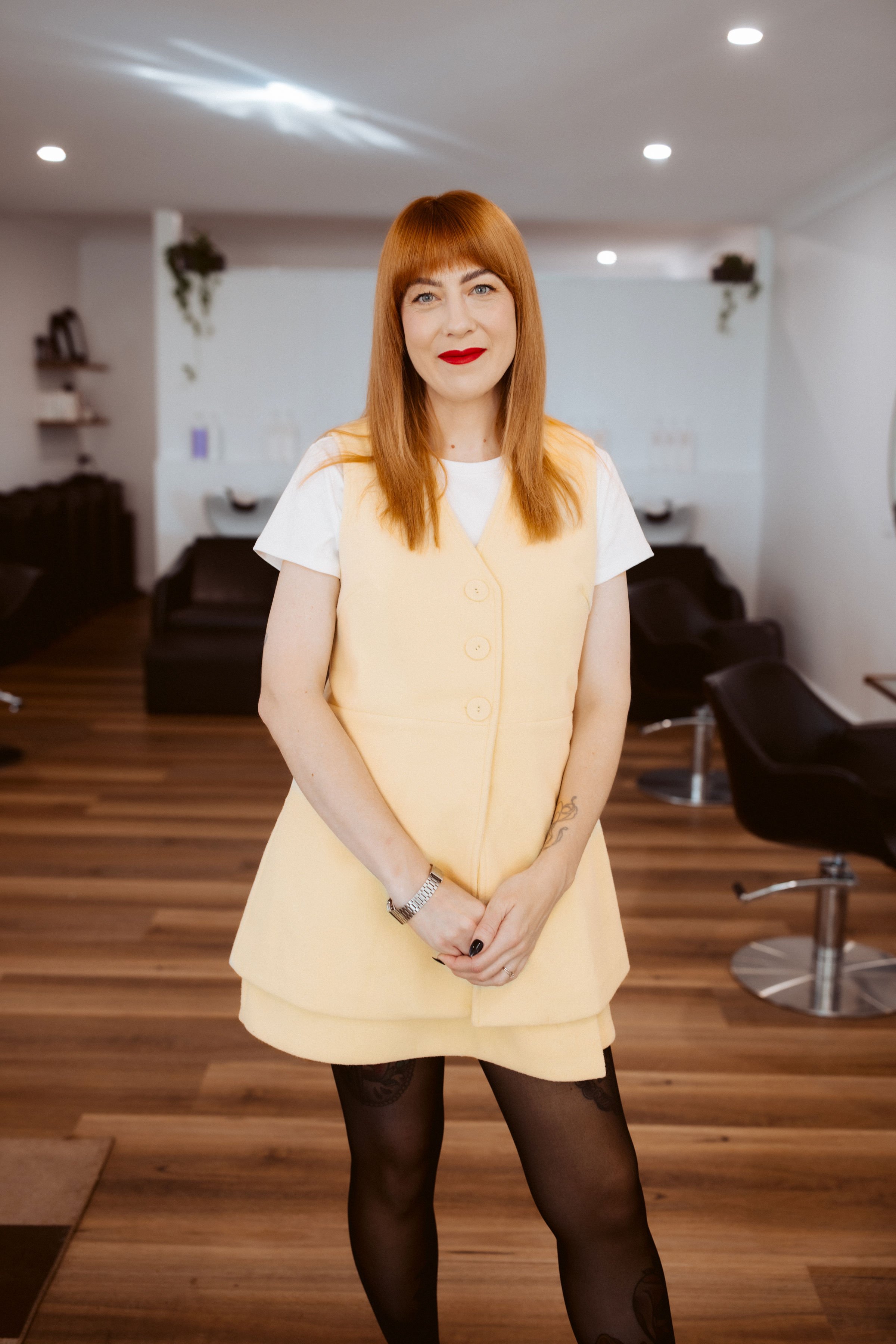 A woman with red hair and red lipstick standing in a hair salon, wearing a yellow vest over a white shirt, black patterned tights, and a watch on her left wrist.