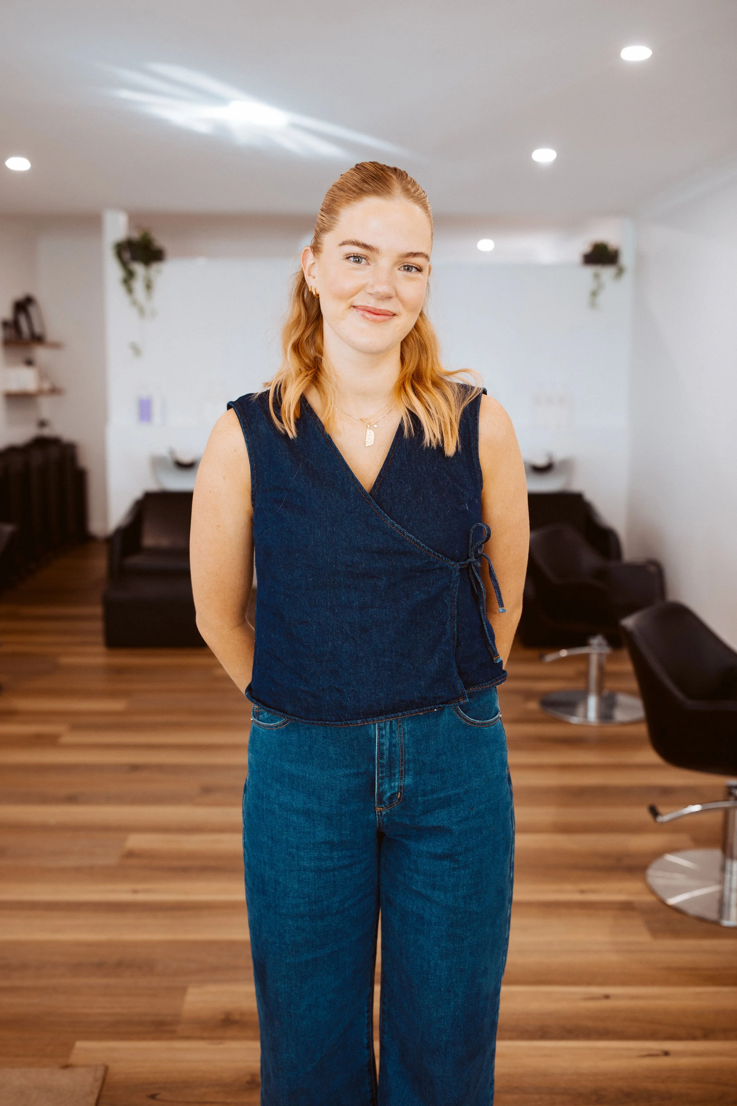 A young woman with red hair, wearing a sleeveless denim top and jeans, standing in a modern hair salon with wooden flooring and salon chairs in the background.