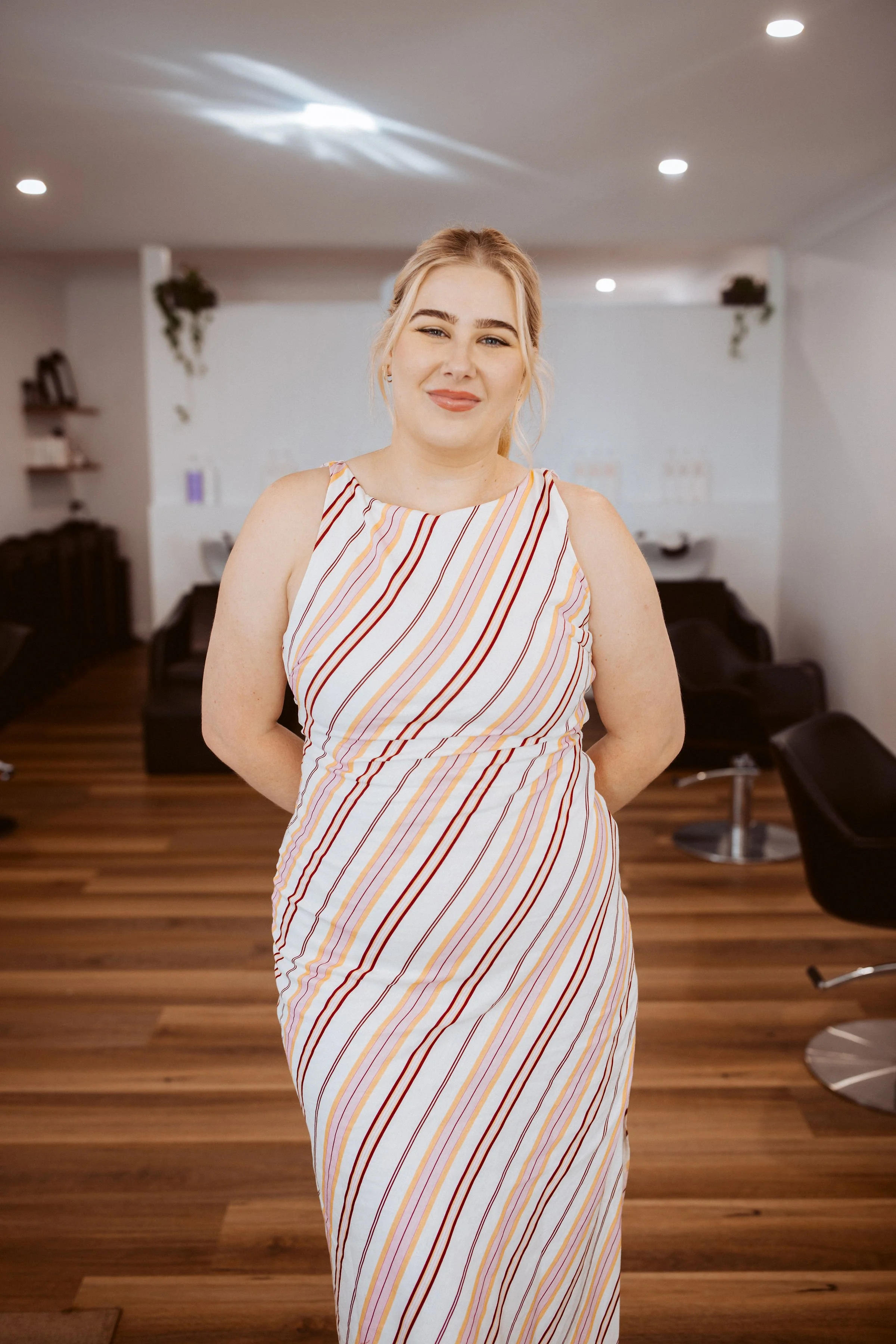 A woman standing in a hair salon, wearing a sleeveless, striped dress with her hands behind her back, smiling at the camera.