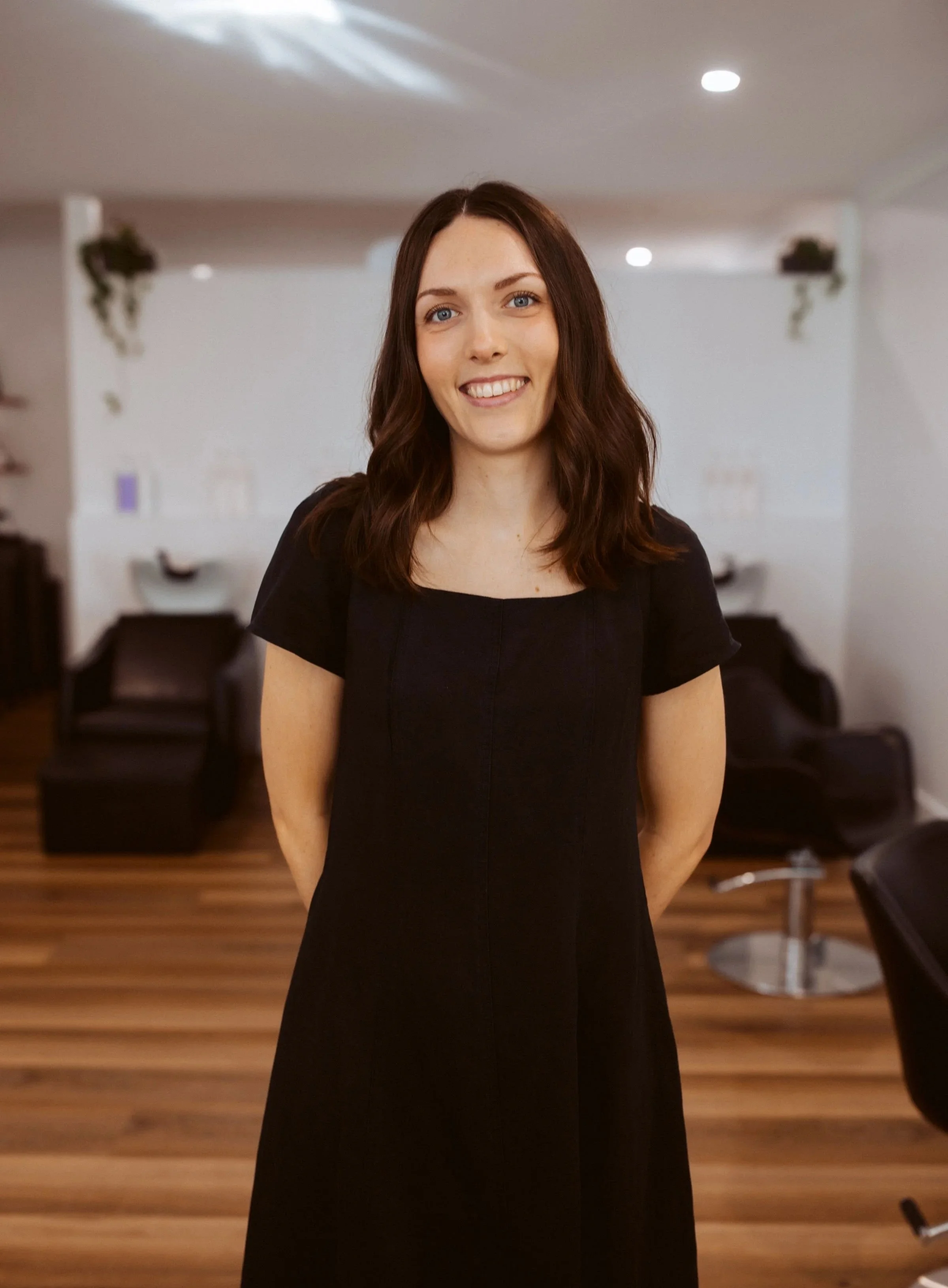 A woman with shoulder-length brown hair, wearing a black dress, standing in a salon with salon chairs and hair washing stations in the background.