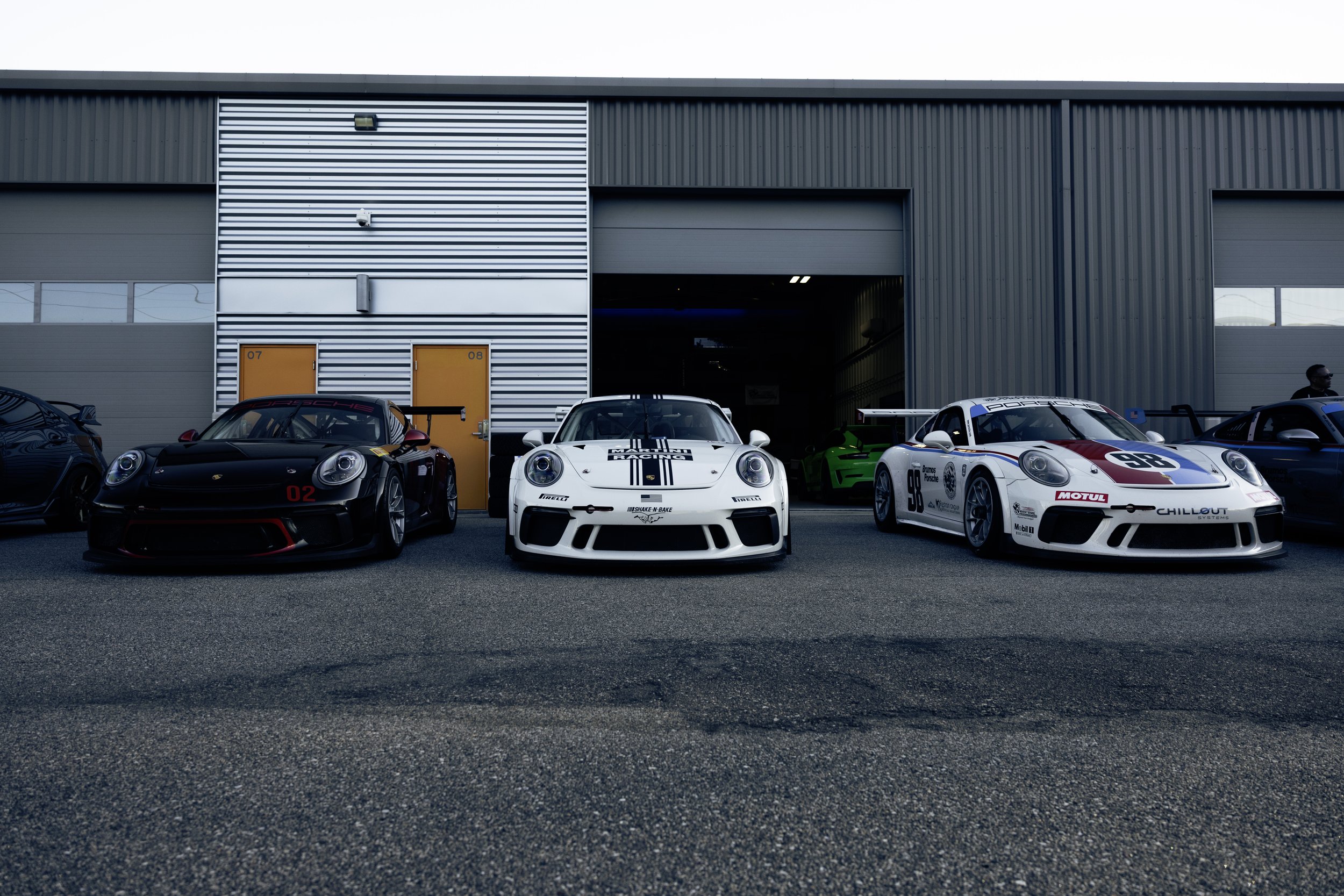 Line-up of three race cars parked outside a garage, featuring a black sports car on the left, a white race car in the middle, and a white race car with red and blue accents on the right, with additional cars visible in the background.