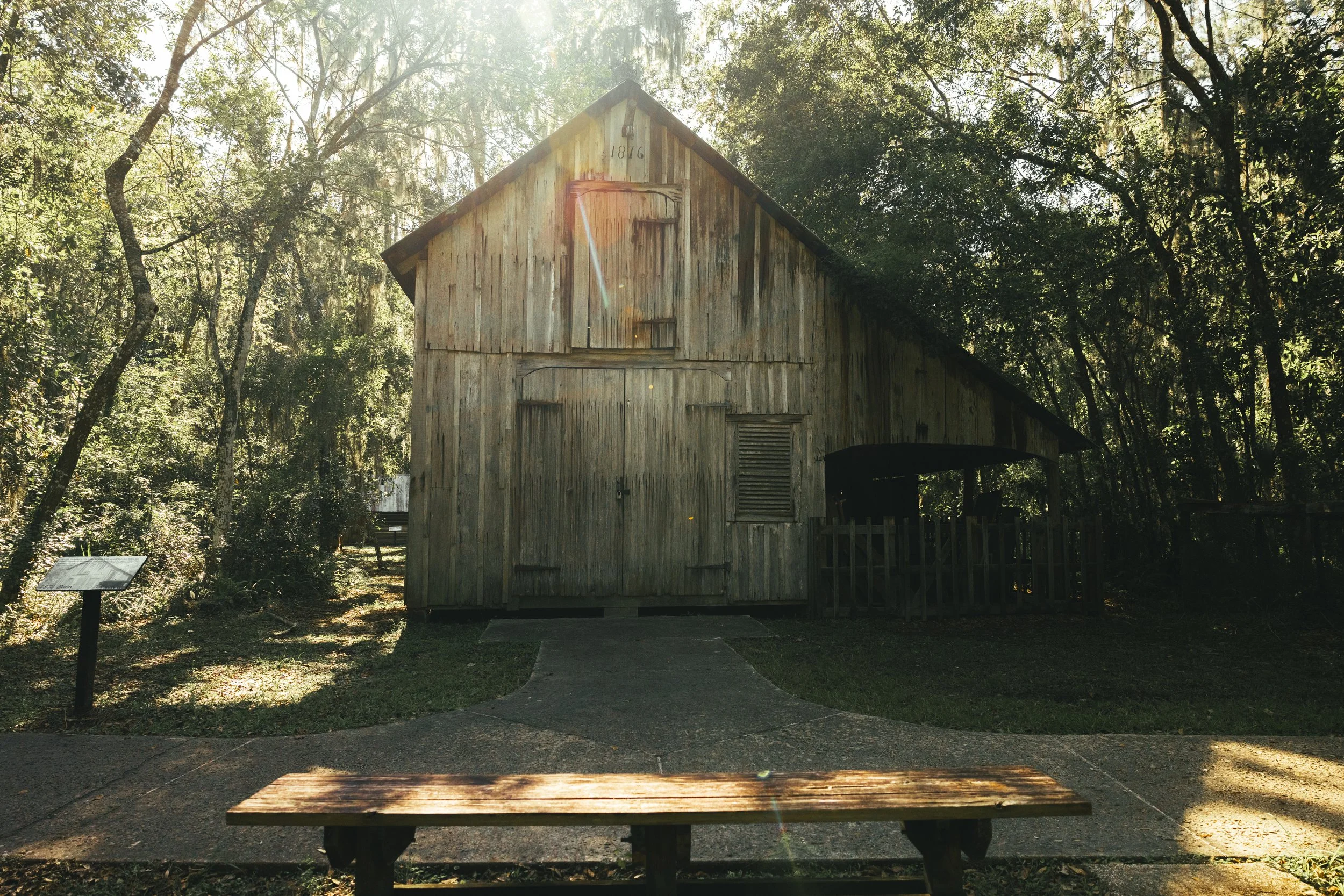 An old wooden barn with weathered planks and a sloped roof, surrounded by trees and greenery, with sunlight filtering through the branches.