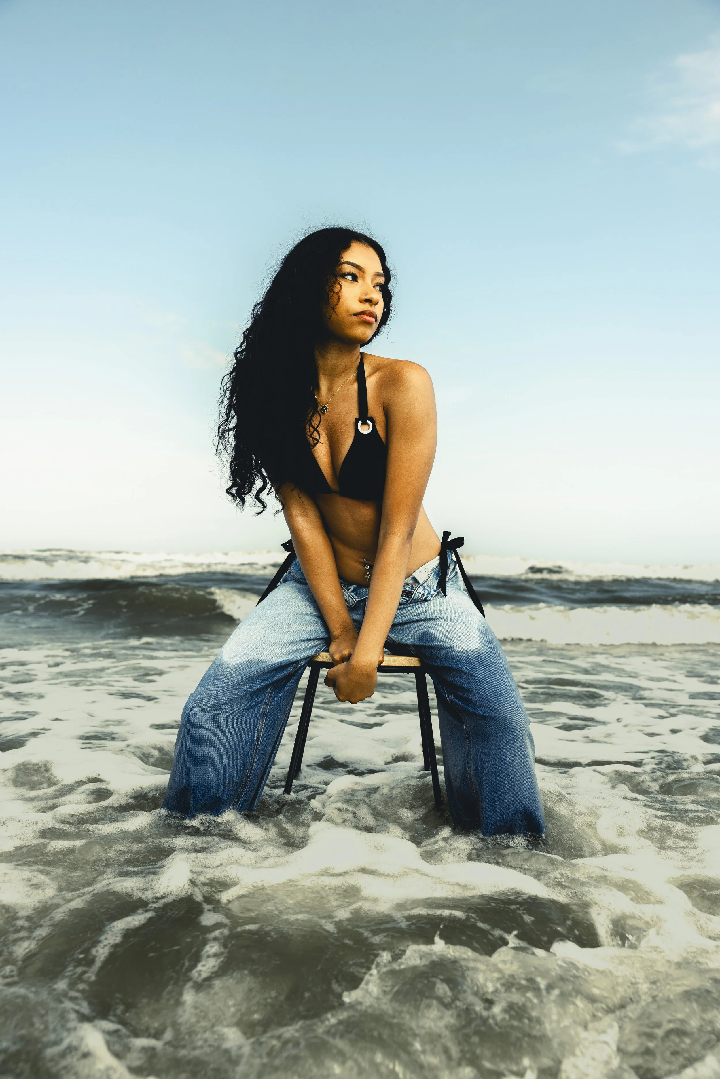 A woman with long black curly hair sitting on a chair in the ocean waves at the beach, wearing a black bikini top and loose blue jeans.