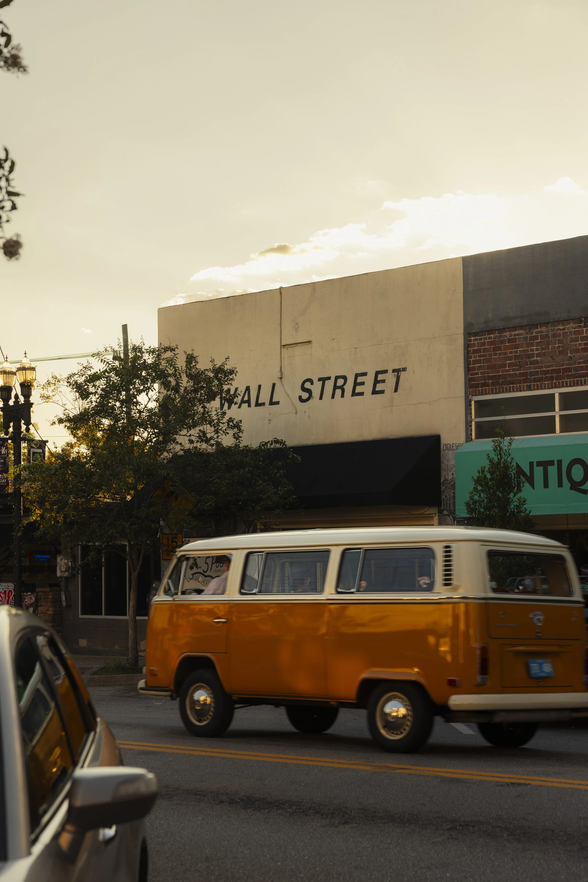 A vintage orange and white Volkswagen van driving past Wall Street storefront at sunset.
