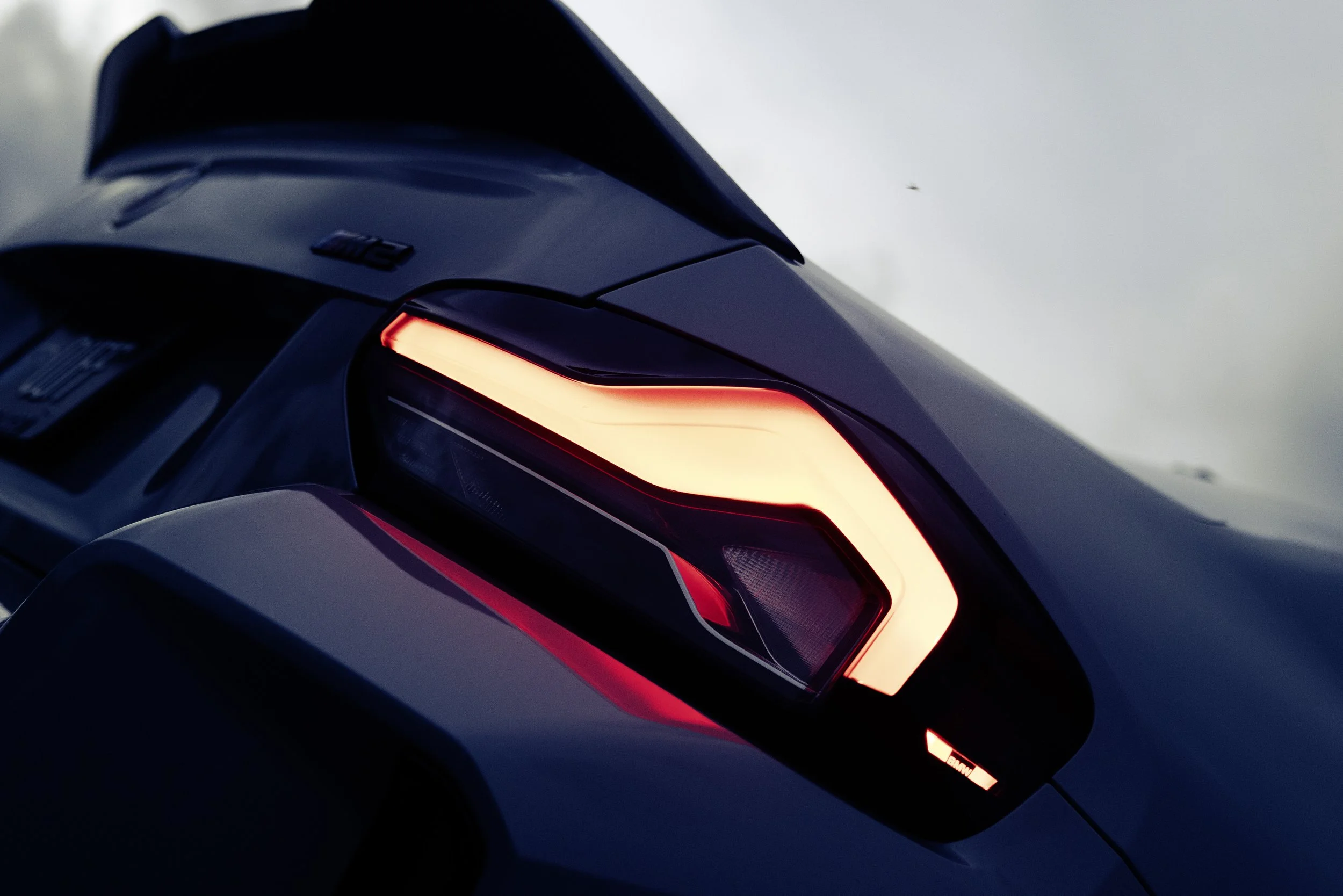 Close-up of a rear car taillight with turn signal illuminated, part of a dark-colored car, with a blurred background of sky.