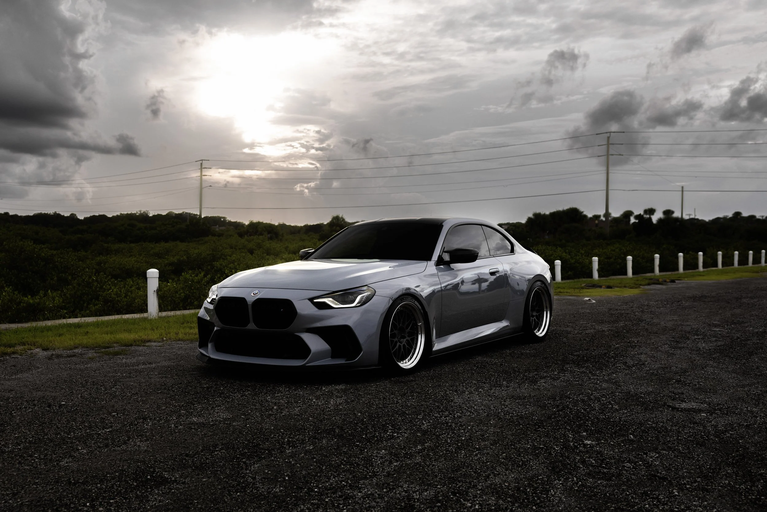 A modern gray sports car parked on a paved roadside with greenery and a cloudy sky in the background.