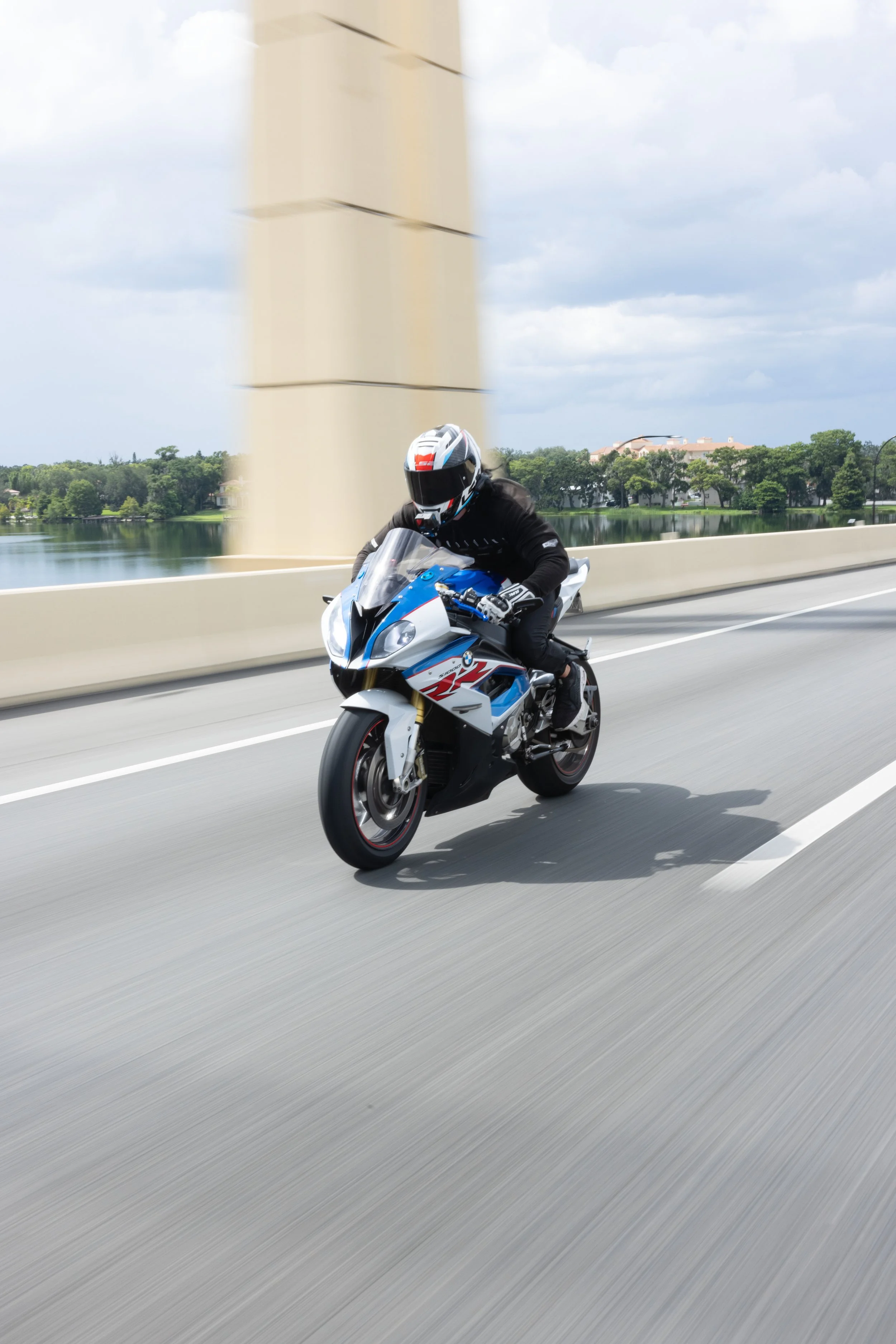 A motorcyclist wearing a black jacket, black pants, and a white helmet rides a blue, white, and black sport bike on a highway with a blurred background of trees, water, and a beige bridge pillar.