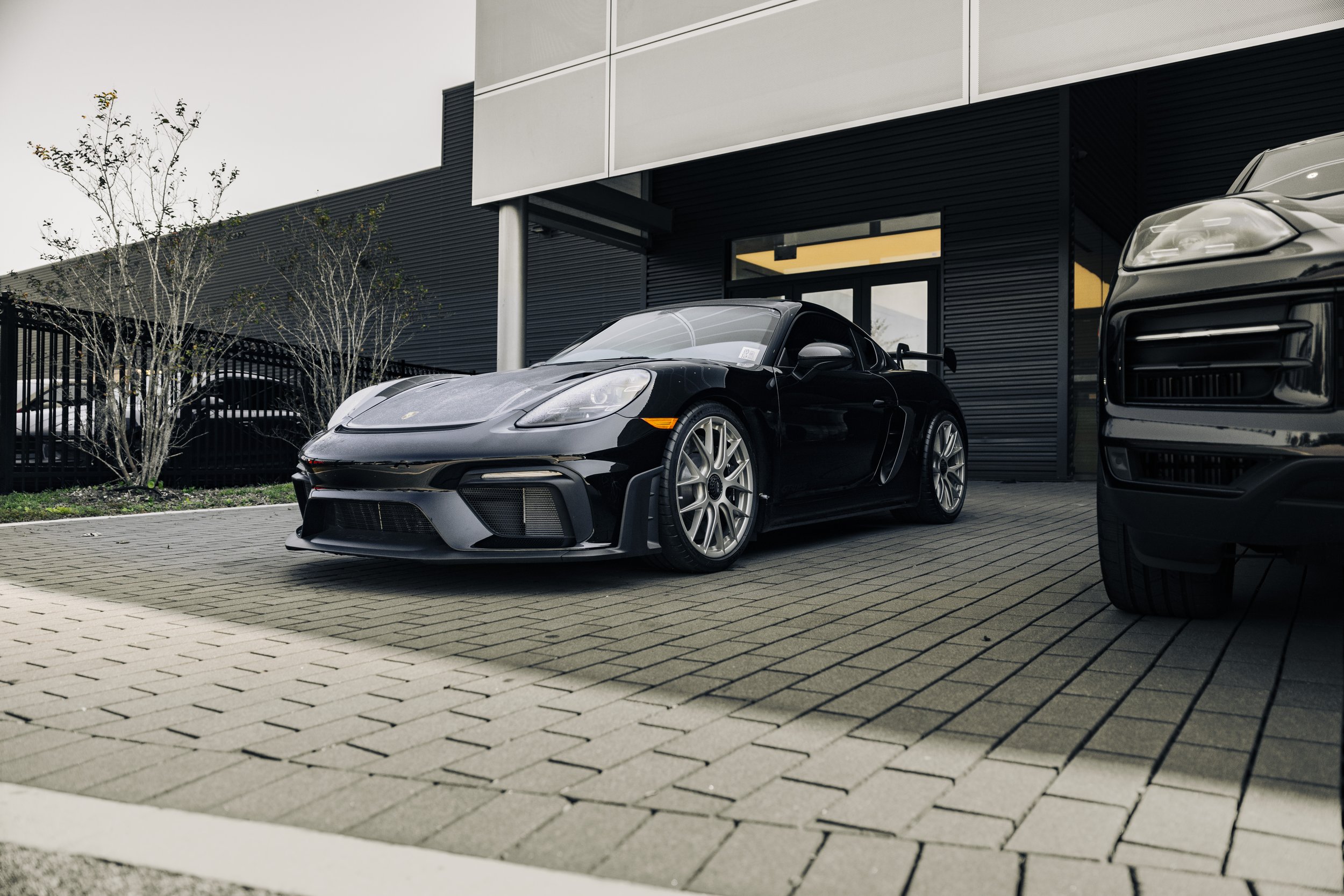 A black sports car parked outside a modern building with black siding and large windows, alongside a black truck on the right, in a paved area with a small tree and fencing in the background.