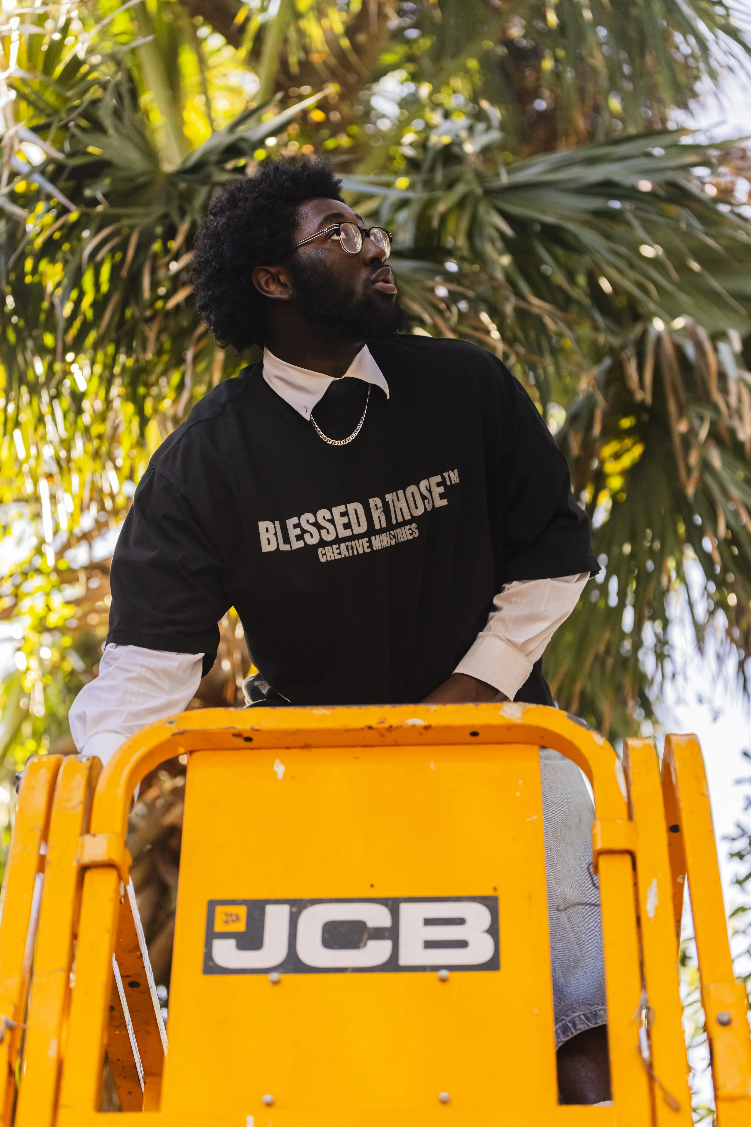 A man with glasses and a beard wearing a black T-shirt with white long sleeves underneath, standing on a yellow JCB lift against a background of leafy green trees.