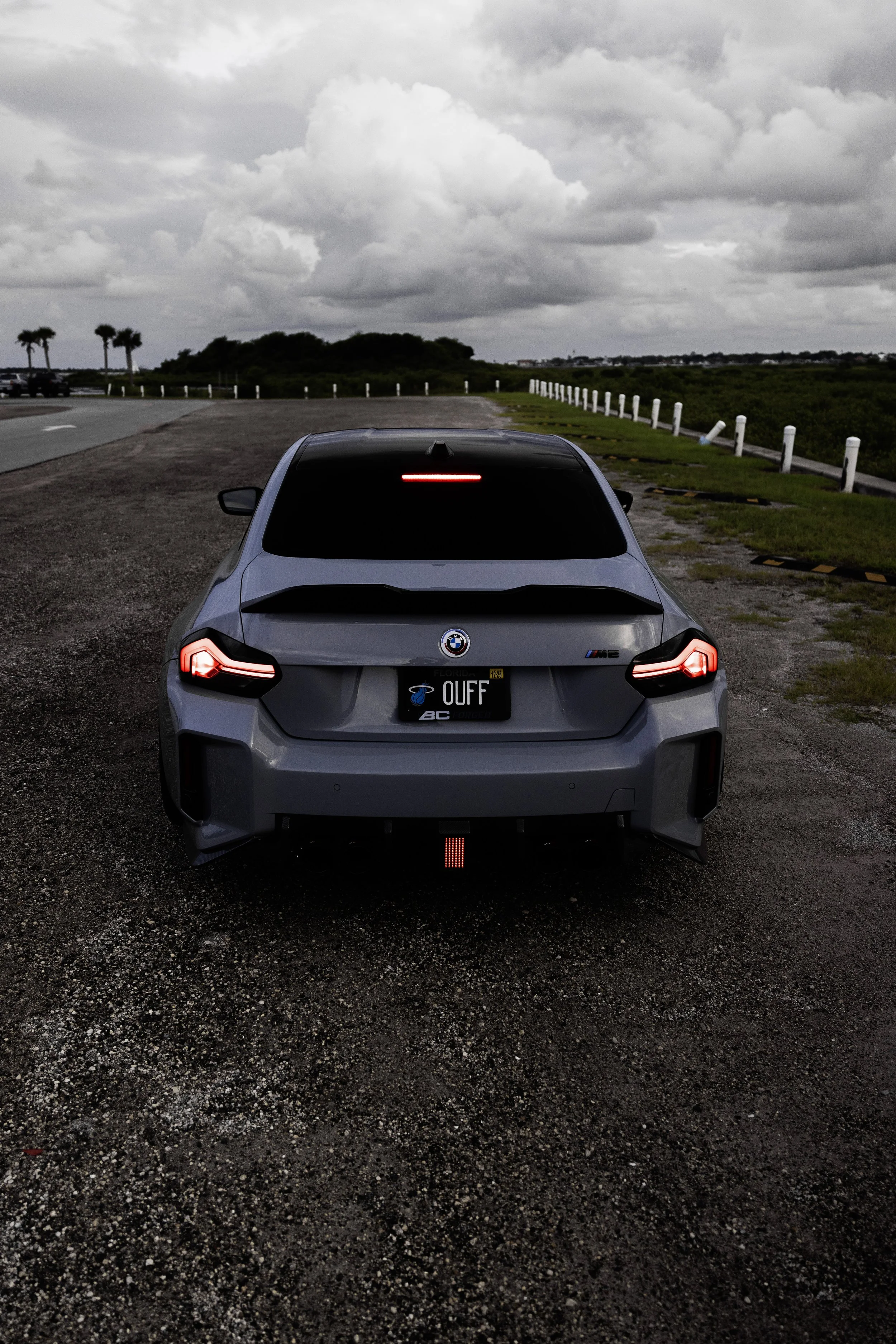 Back view of a gray BMW SUV parked on gravel with a sports-themed license plate that reads 'OUFF' and a red safety light on the rear. Overcast sky with large clouds in the background.