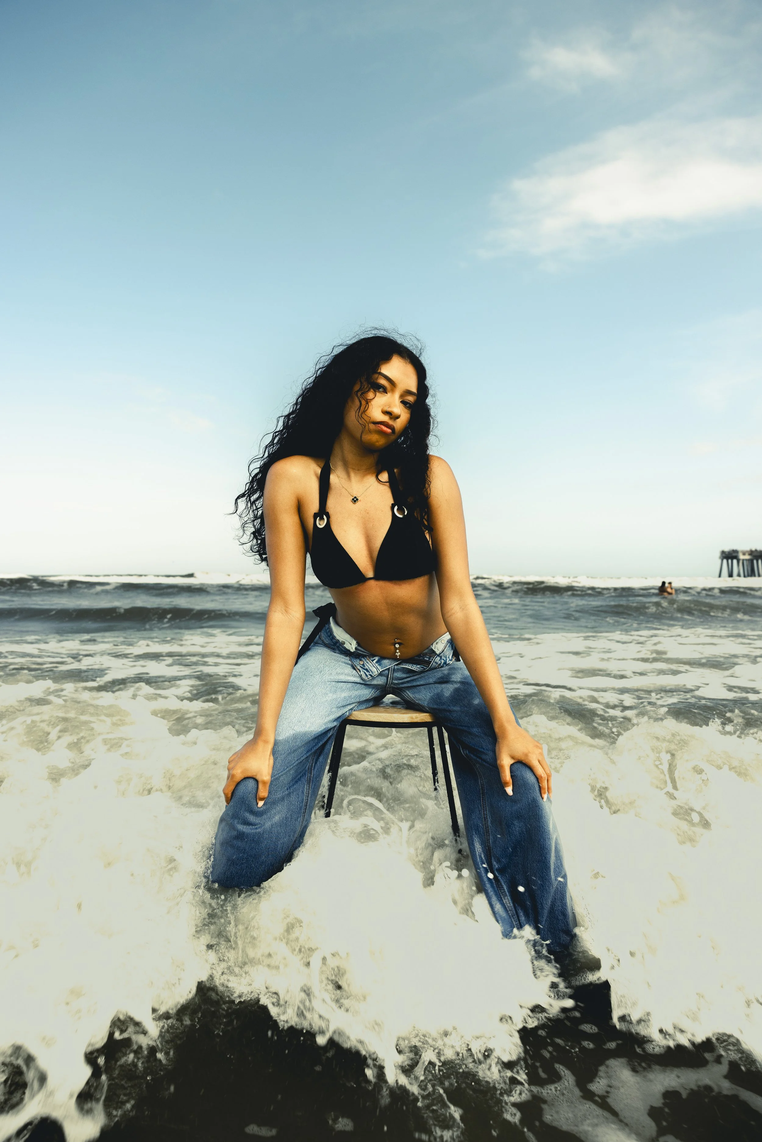 A young woman with curly black hair, wearing a black bikini top and loose blue jeans, sitting on a stool in the ocean with waves around her, looking at the camera against a partly cloudy sky with a pier in the distance.