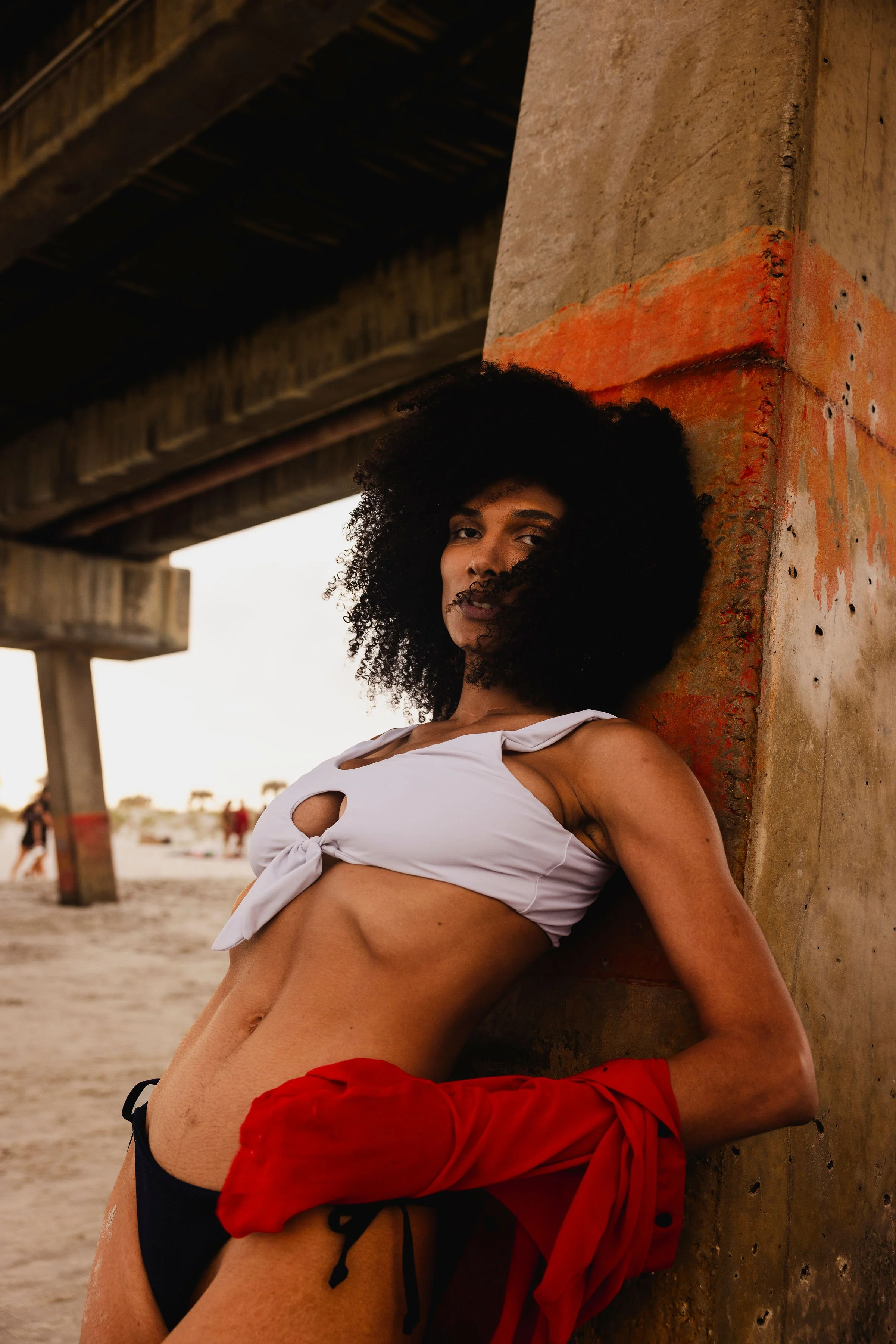 A woman with curly black hair leaning against a concrete pillar under a bridge, wearing a white tied crop top, black underwear, and red sleeves tied around her waist, on a beach with other people in the background.
