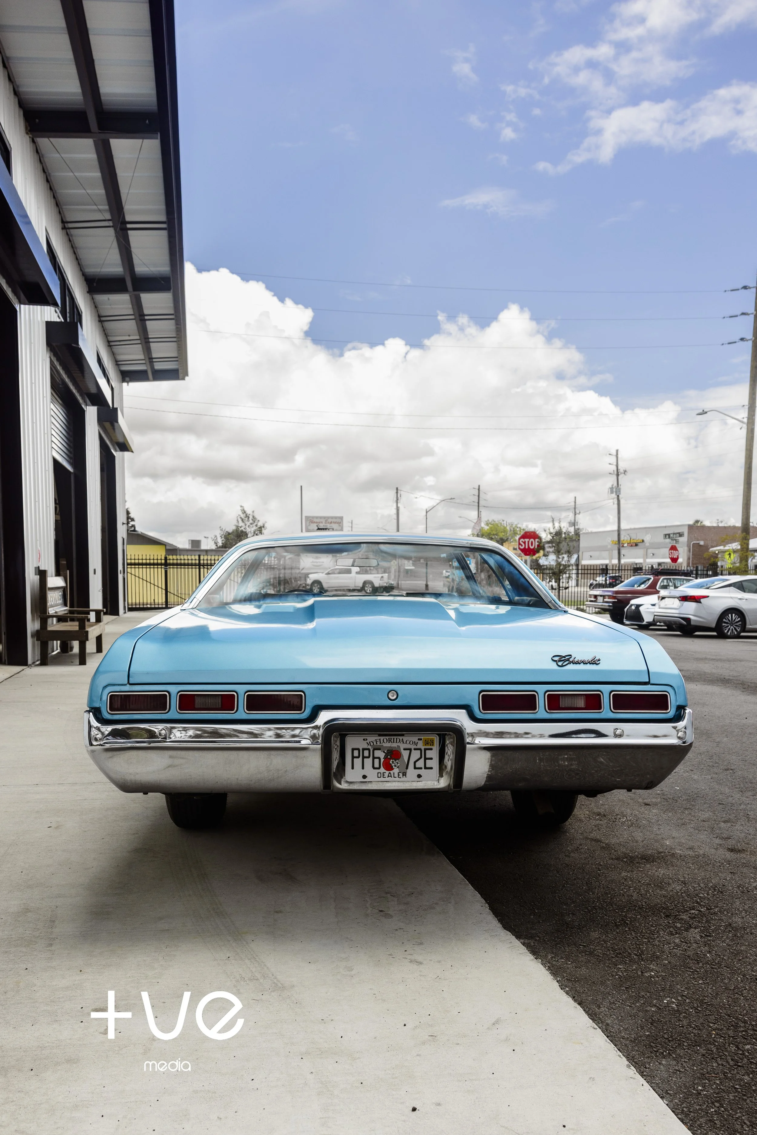A light blue vintage Chevrolet car parked on a concrete sidewalk, with the rear facing the camera, under a partly cloudy sky.