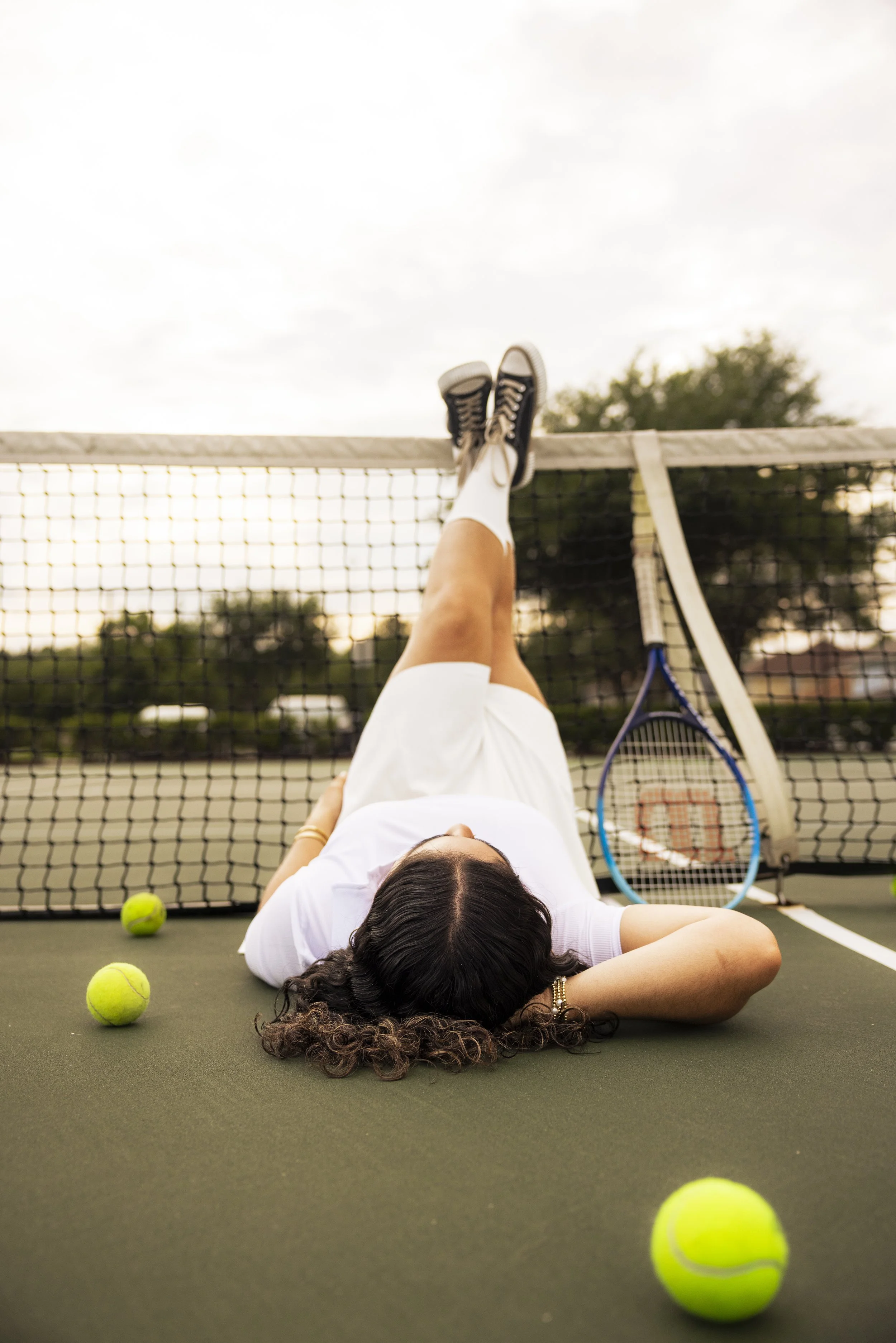 A woman lies on a tennis court with her legs propped up on the net, surrounded by tennis balls, with a tennis racket resting against the net.