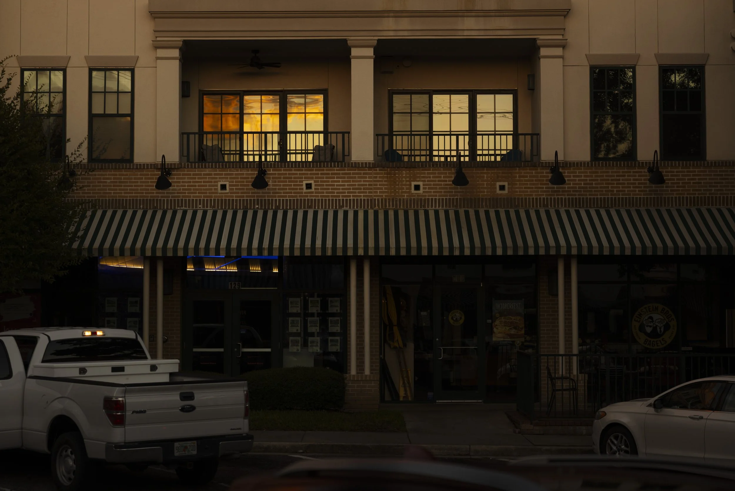 A two-story building with a striped awning on the ground floor and a balcony with chairs on the upper floor. The upper windows reflect the sunset sky. Parked cars are visible in front of the building.