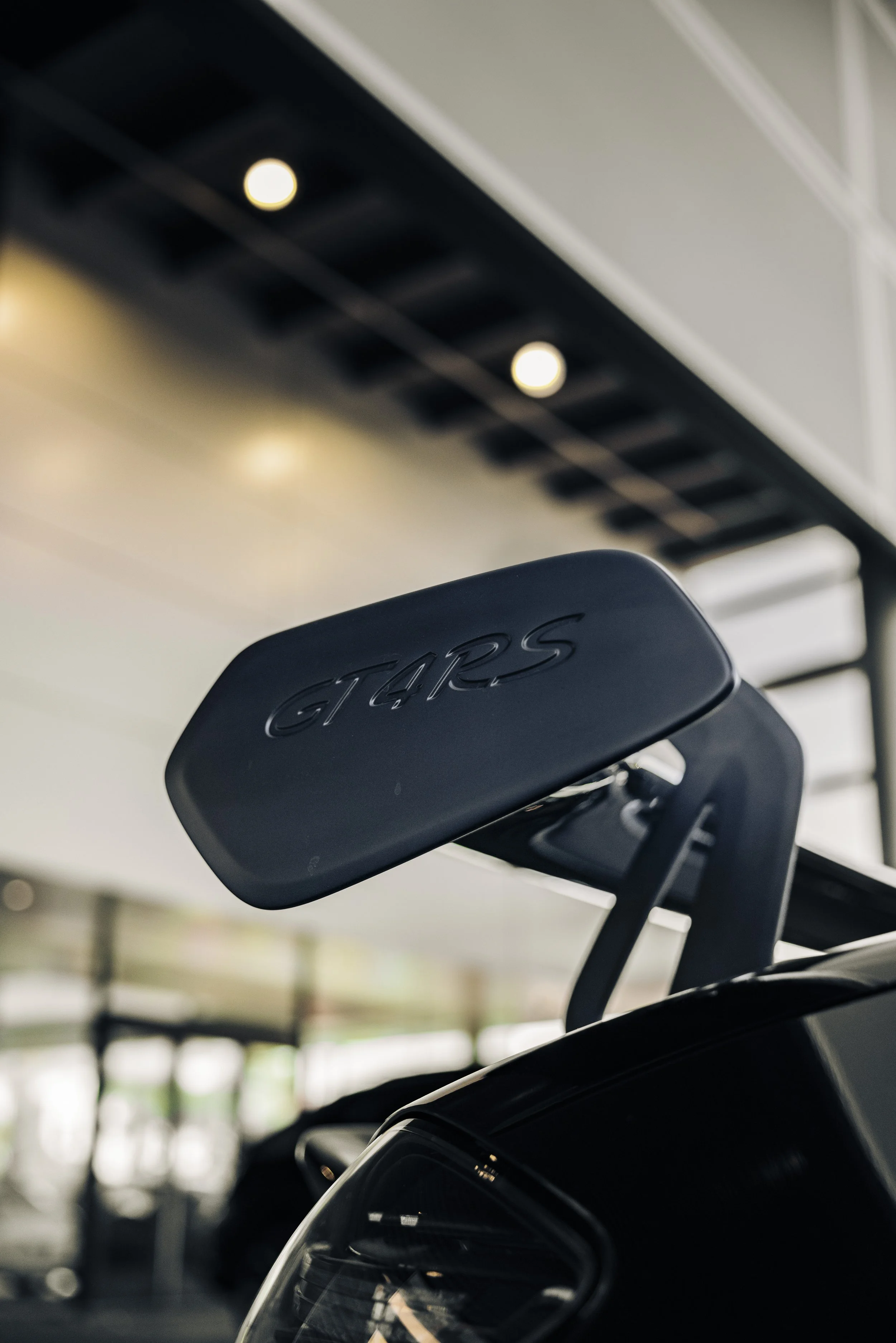 Close-up of a black Porsche GT4 car's rear wing with the GT4 logo engraved, in an indoor showroom or garage.