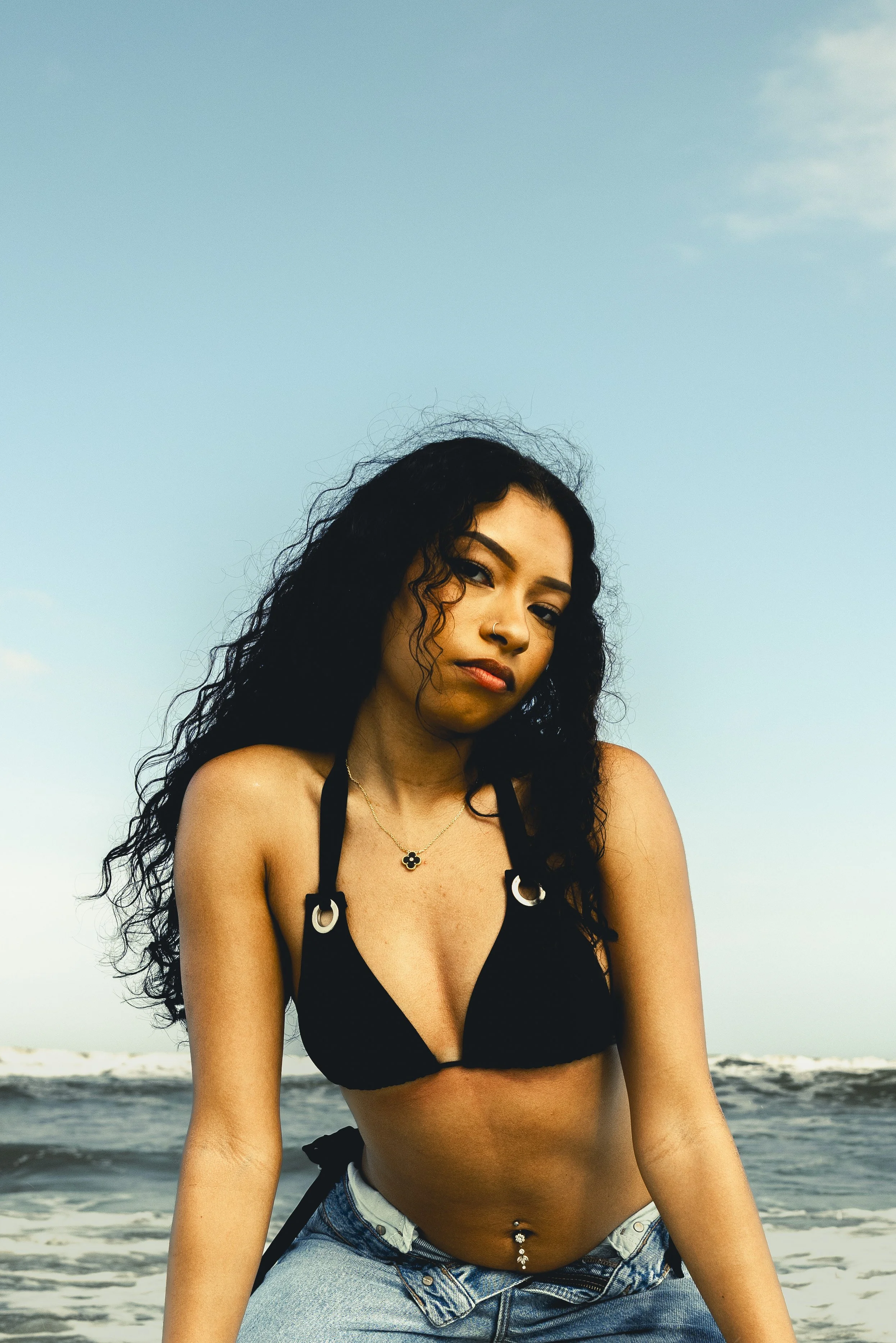A woman with curly black hair and tan skin posing on a beach. She is wearing a black bikini top with metallic ring details and light blue jeans, with a navel piercing. The sky is clear with some clouds, and the ocean is in the background.