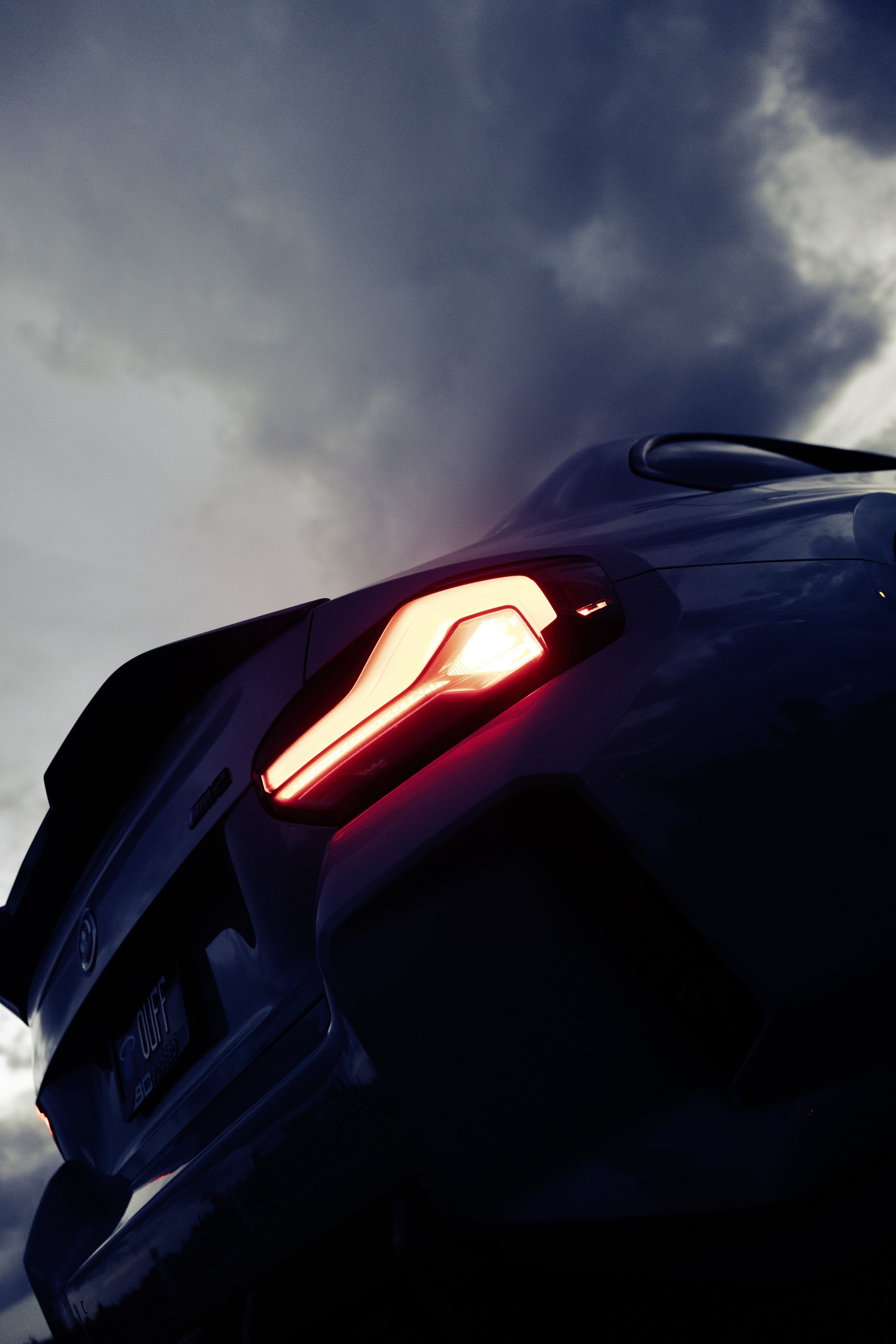 The rear view of a blue BMW M3 sports car with illuminated tail lights under a cloudy sky.