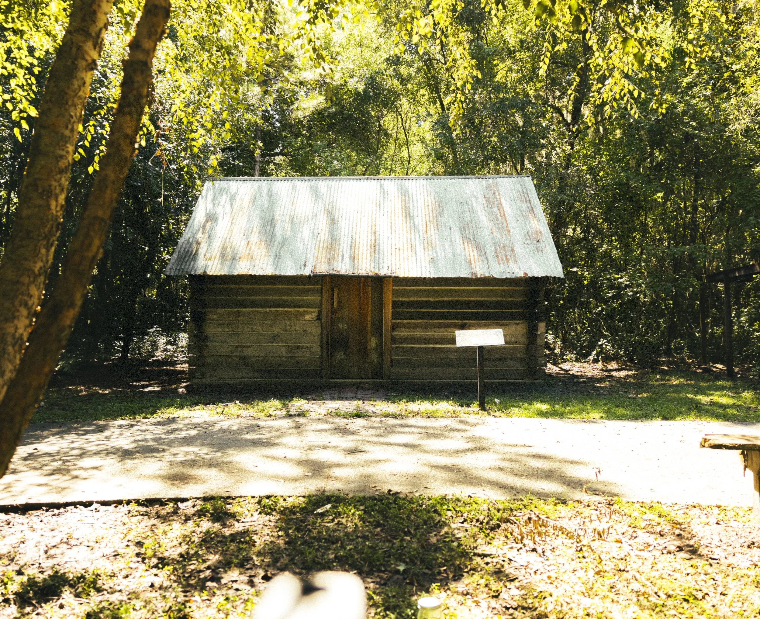 A small wooden cabin with a rusted metal roof is situated in a wooded area, with a signpost in front of it and trees surrounding the scene.
