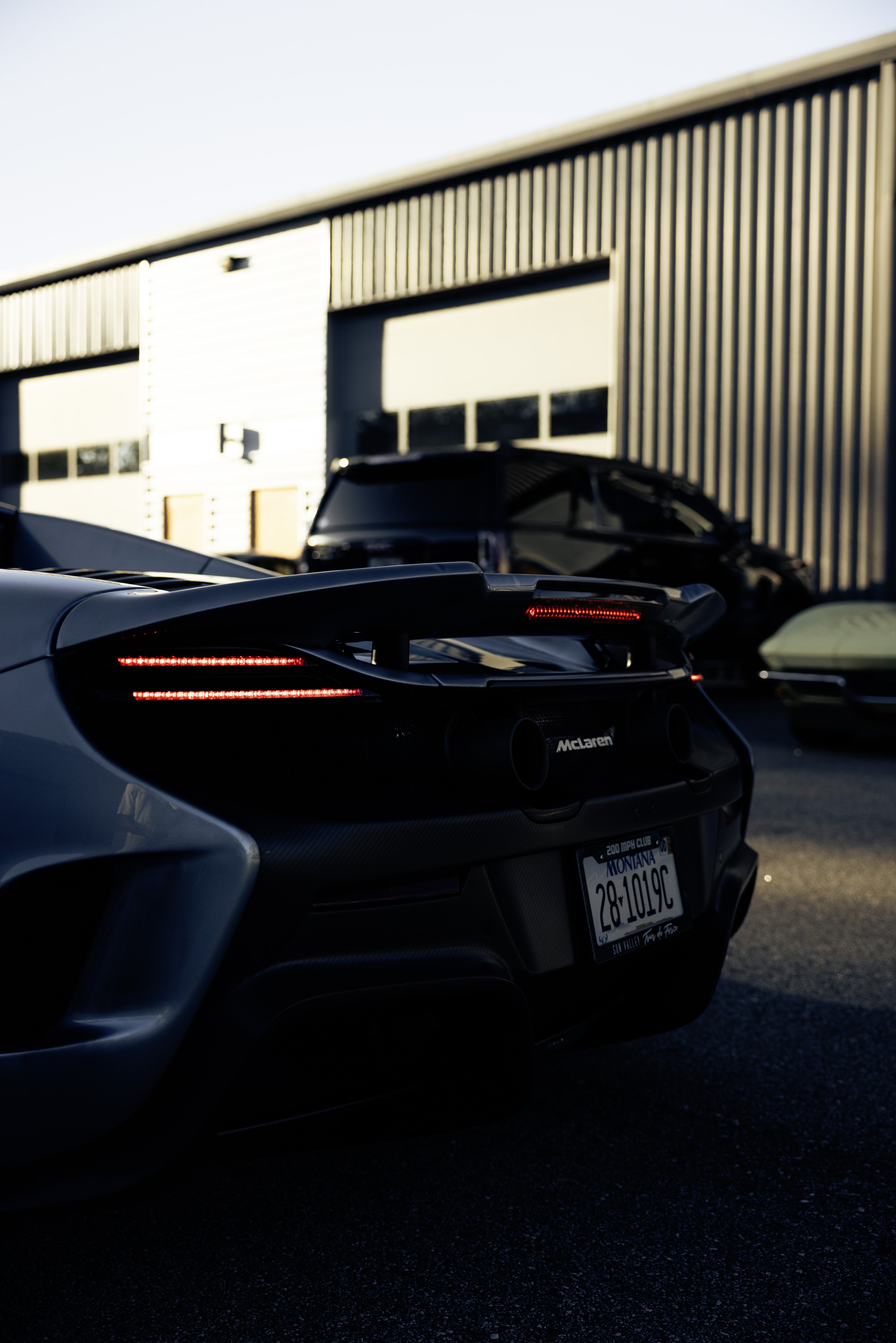 Rear view of a silver McLaren sports car with a Montanan license plate, parked outside a building with a metal exterior, during sunset.