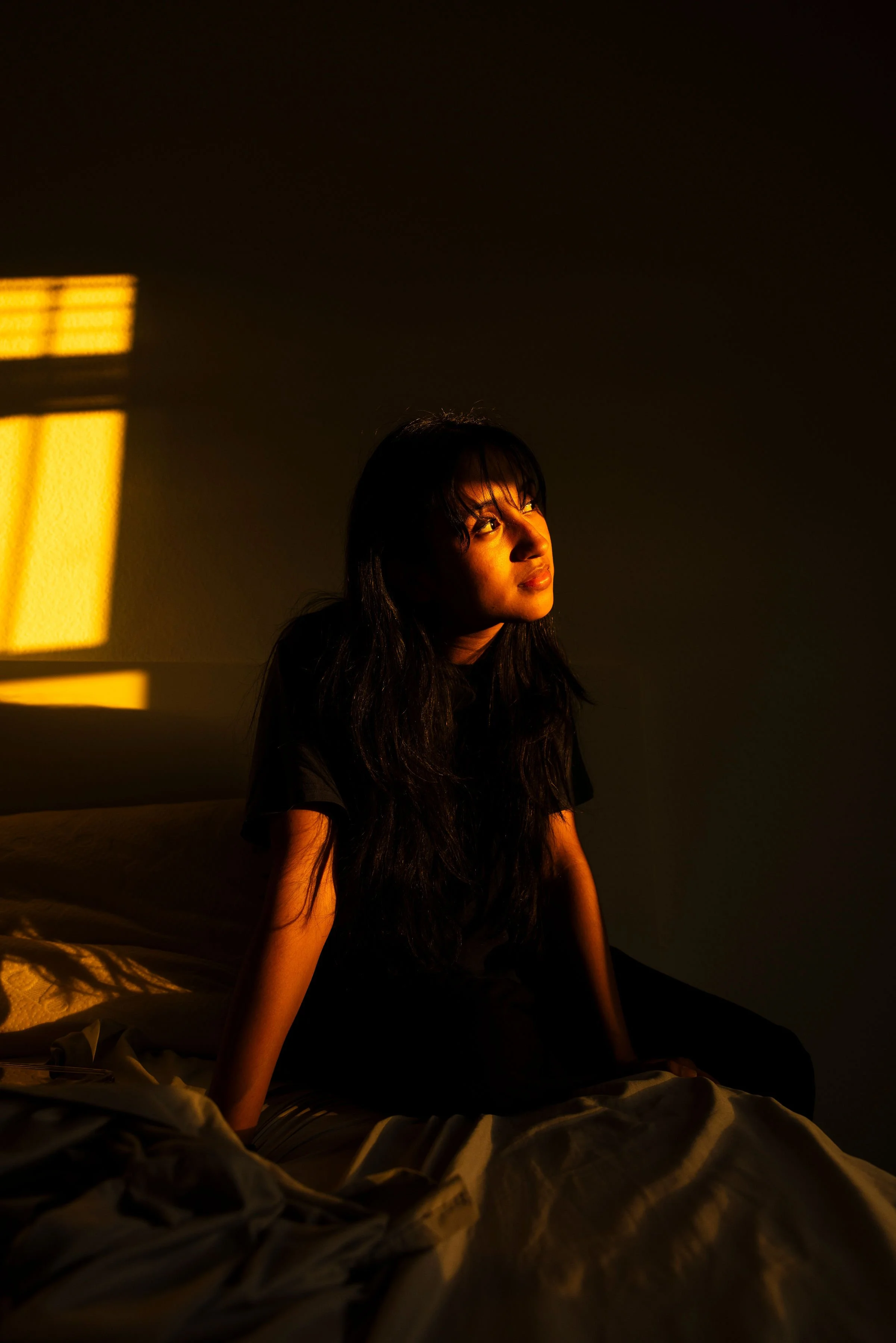 A woman with long black hair and bangs sitting on a bed in a dimly lit room, illuminated by warm sunlight casting shadows on the wall.