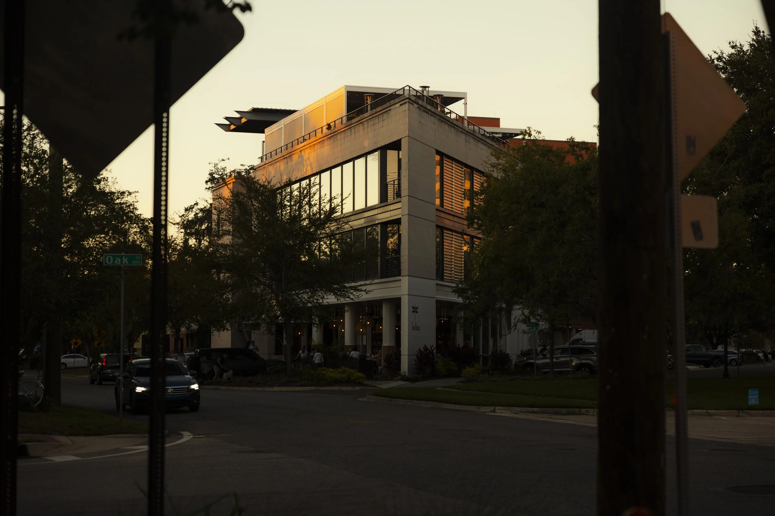 Modern multi-story building at sunset with reflective glass windows, trees in the foreground, street signs, cars, and a few pedestrians.