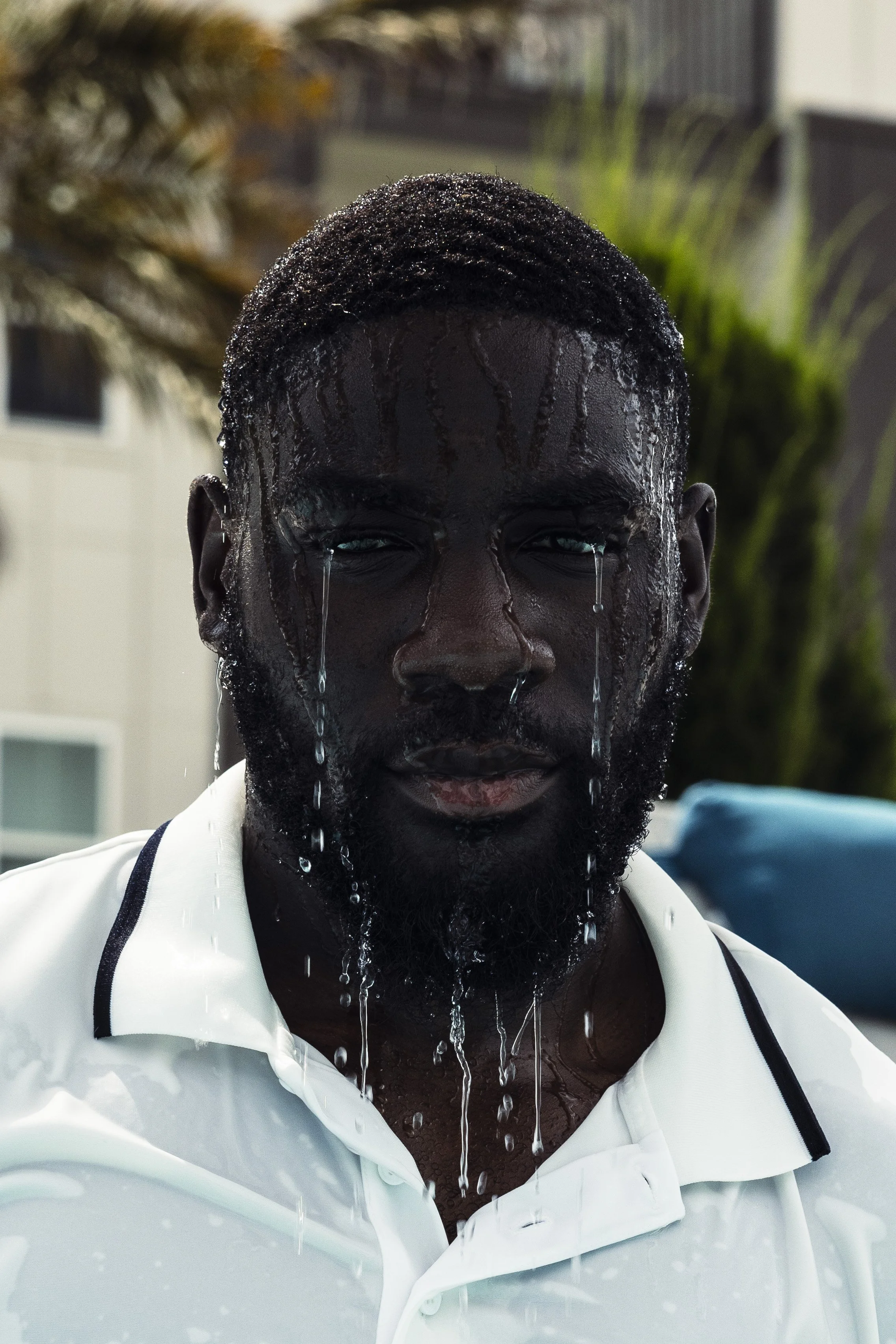 A man with water dripping down his face and hair, wearing a white collared shirt with black trim, outdoors with blurred background of trees and buildings.