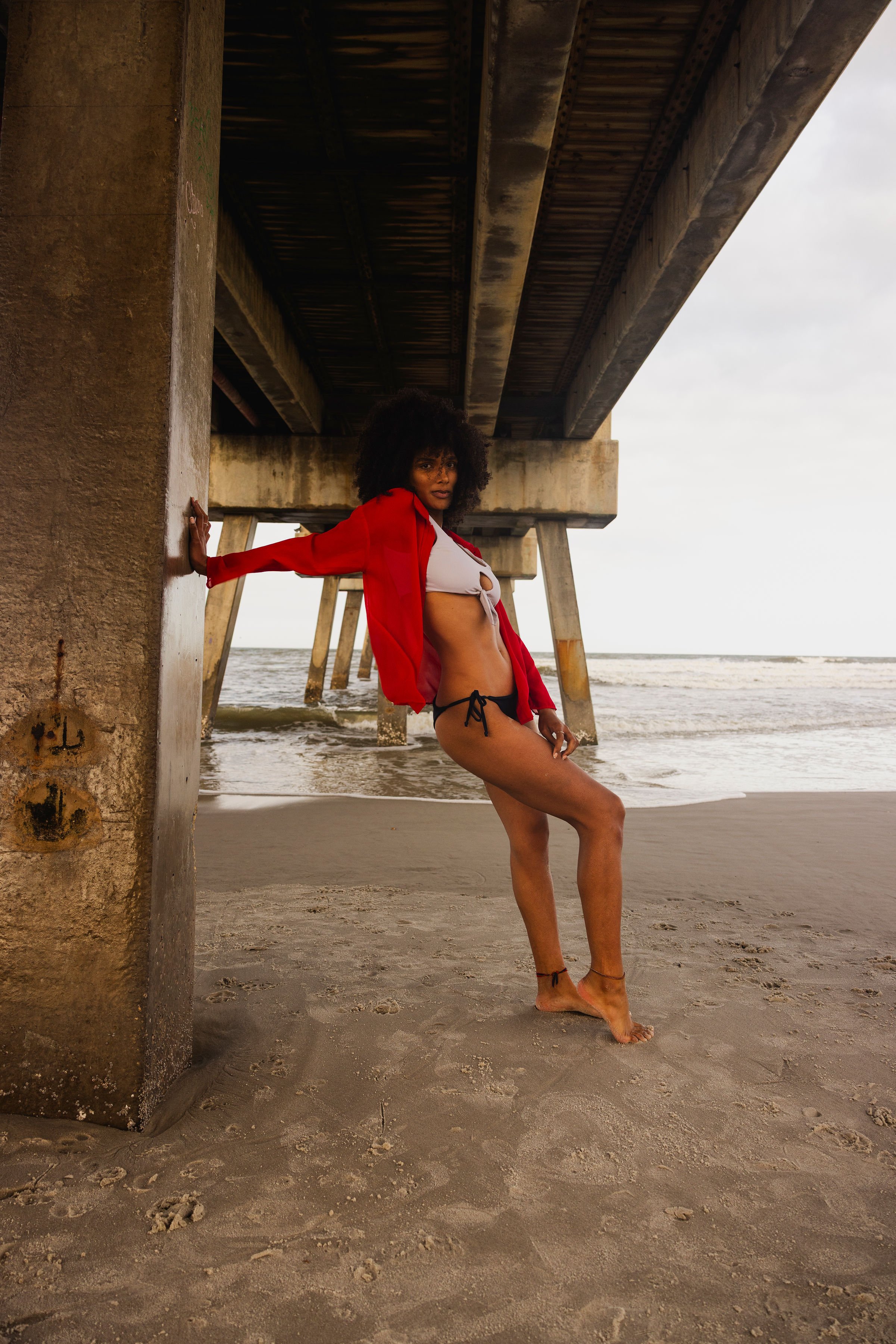 A woman with curly hair in a bikini and red cover-up standing under a pier on the beach, with her arm resting against a concrete pillar and facing the camera.
