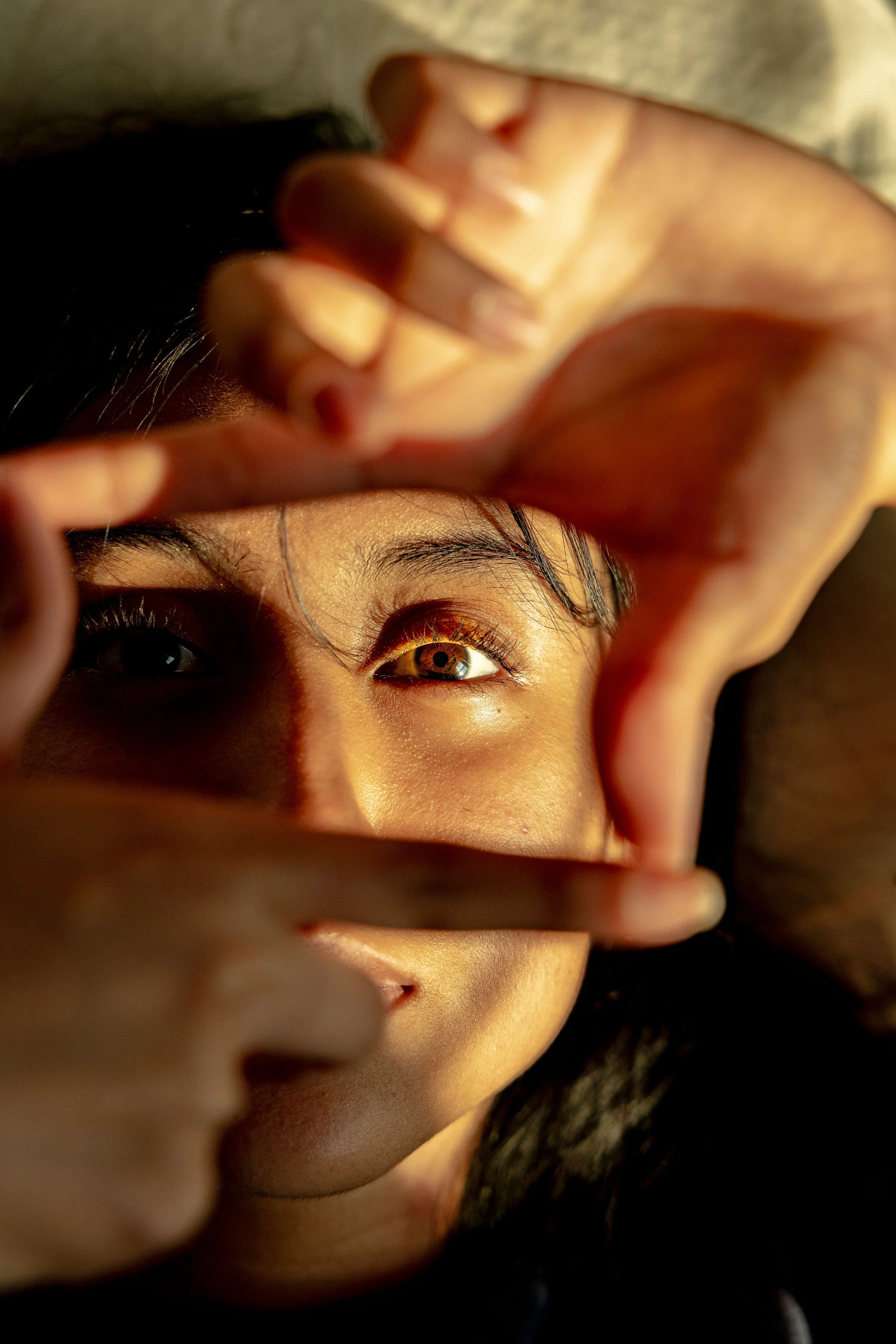 A person holding a mirror in front of their face, showing one eye with a golden-brown iris and light reflecting on the skin near the eye.