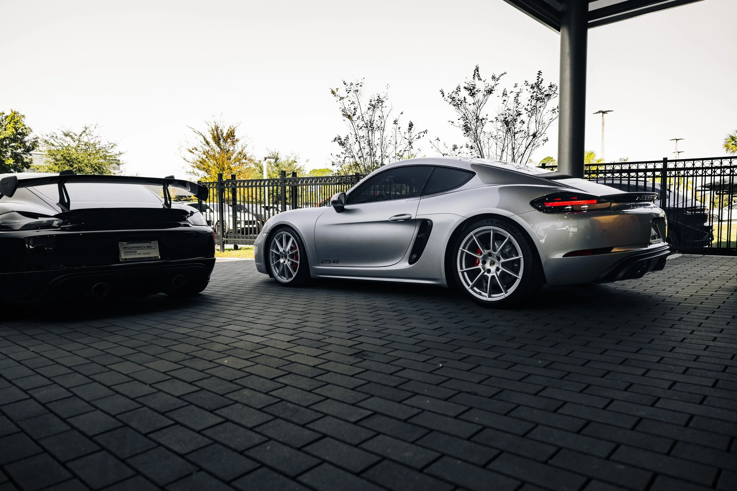 Silver Porsche 718 GTS 4.0 sports car parked next to a black sports car with a spoiler, in a parking area with a black fence and trees in the background.