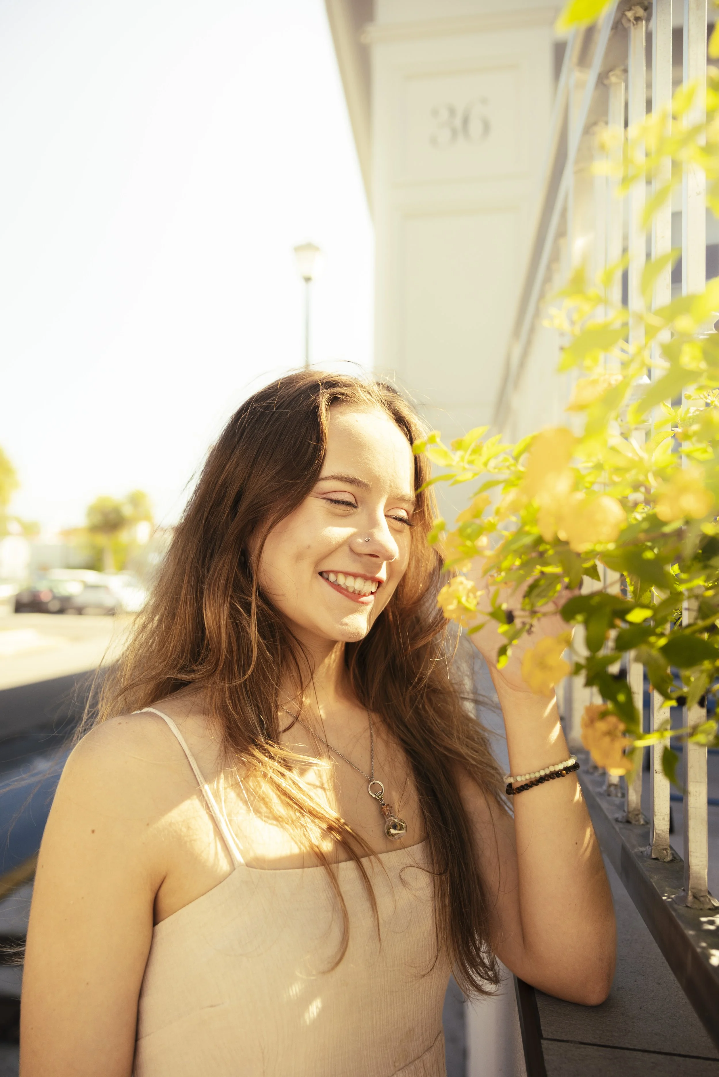 A young woman with long brown hair smiling happily, wearing a beige sleeveless top, standing outdoors near a white fence with yellow flowers in her hand, illuminated by sunny weather.