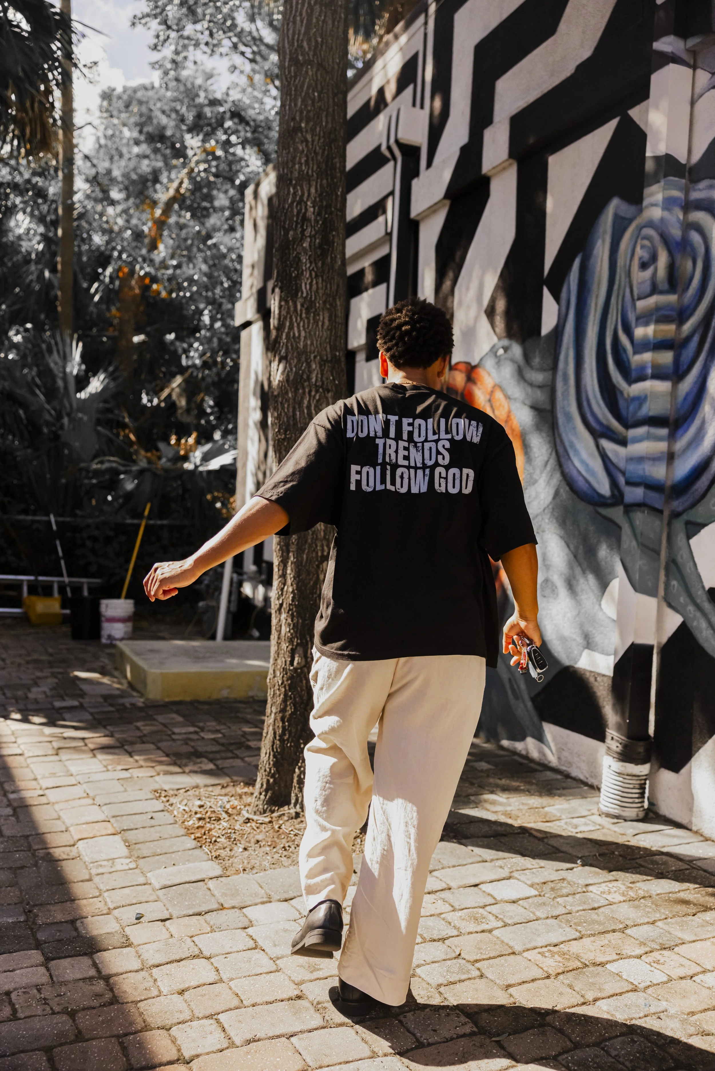 A person wearing a black T-shirt with the phrase "Don't follow trends follow God" on the back, walking on a brick sidewalk near a colorful mural, holding keys in their right hand.
