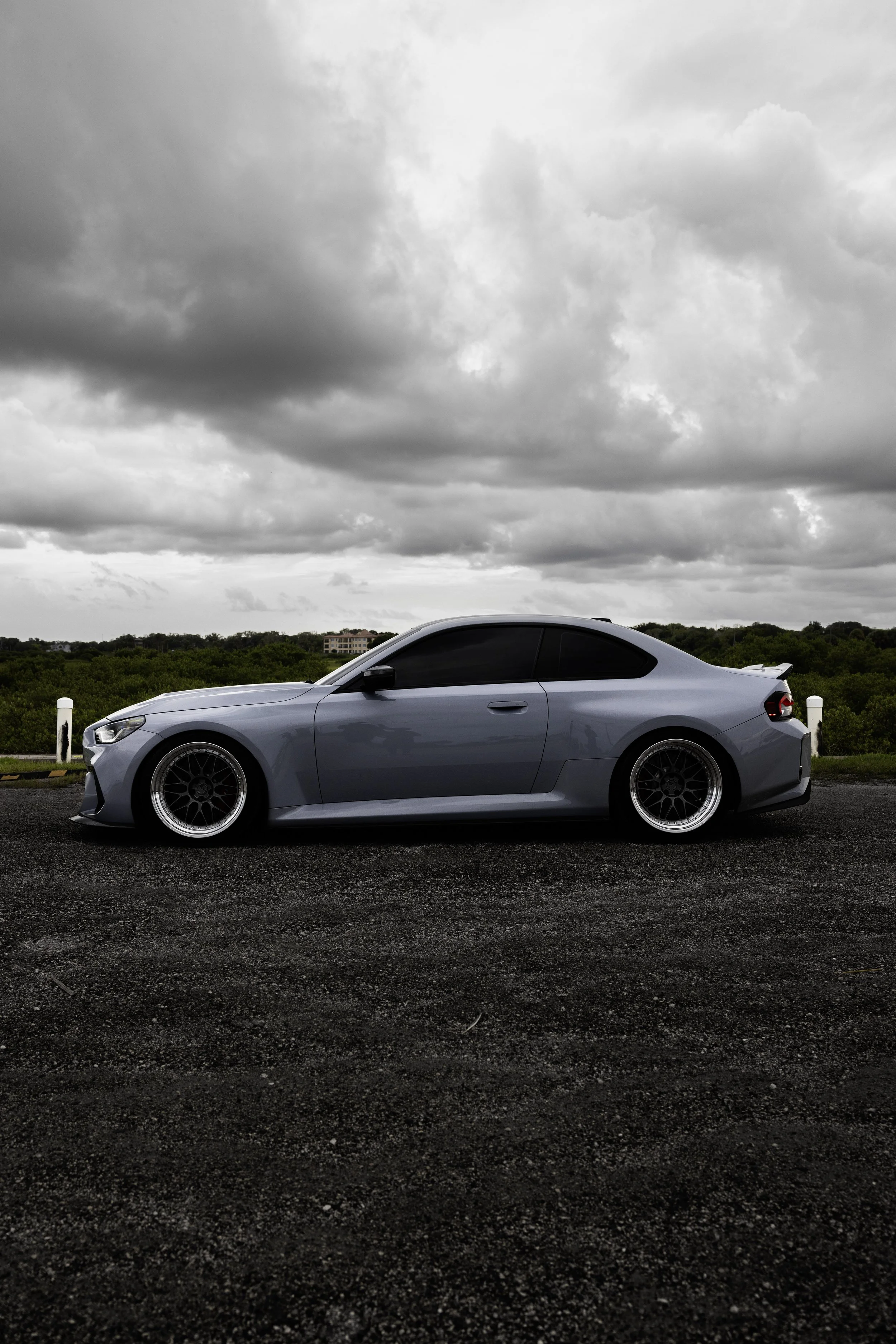 A silver sports car parked on a dark gravel surface under a cloudy sky, with greenery in the background.